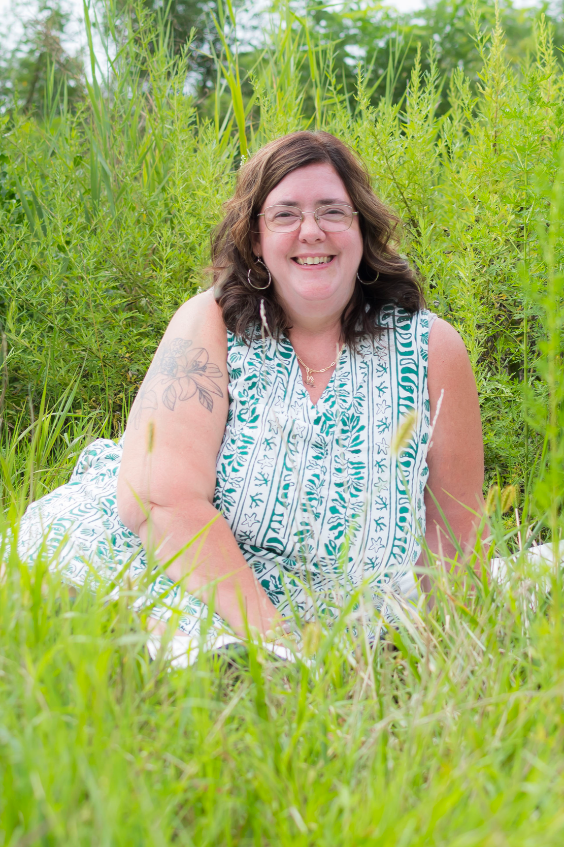 A woman with glasses, tattoos, and dark brown hair smiling while lying in tall green grass surrounded by lush greenery in an outdoor natural setting.
