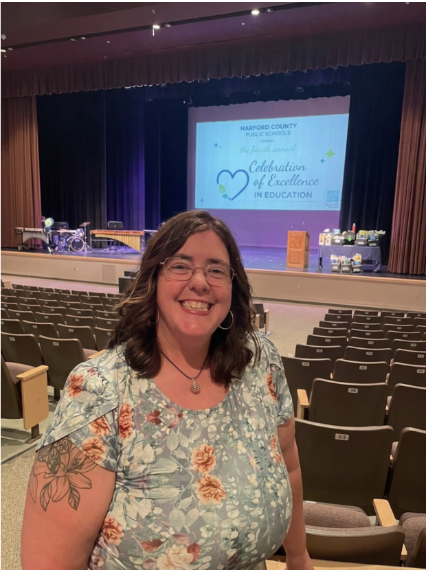 A woman smiling in an auditorium with a stage and a presentation screen in the background. The screen reads 'Harford County Public Schools, the fourth annual, Celebration of Excellence in Education.'