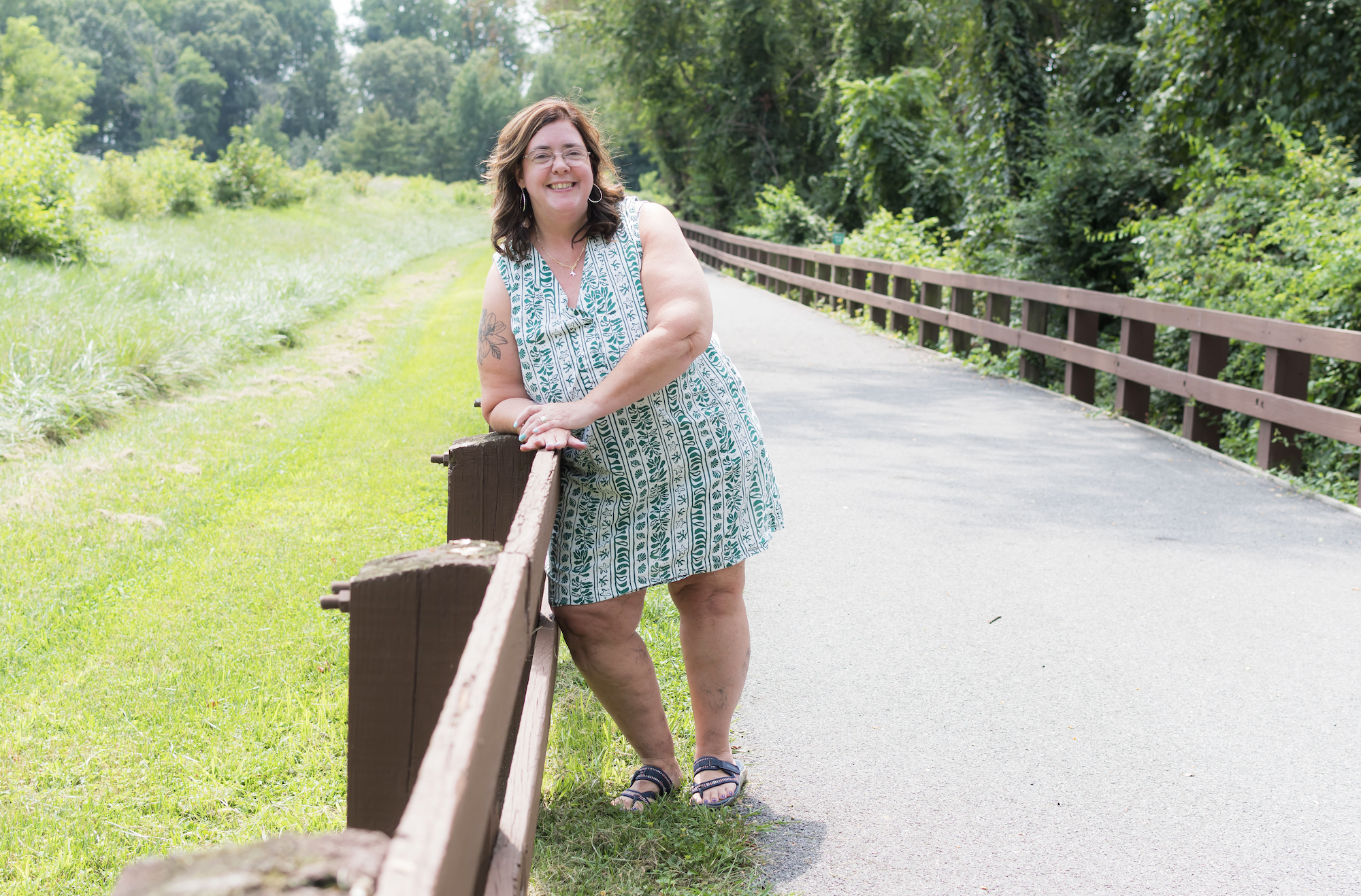 A woman with dark brown hair, glasses, and a tattoo on her left arm, smiling and leaning on a wooden railing on a park trail, surrounded by green trees and grass.
