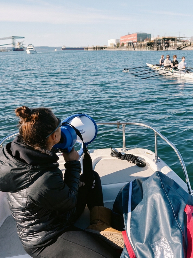 Person with brown hair in a bun looking through a megaphone on a boat, with rowing team in the water ahead and harbor buildings in the distance.