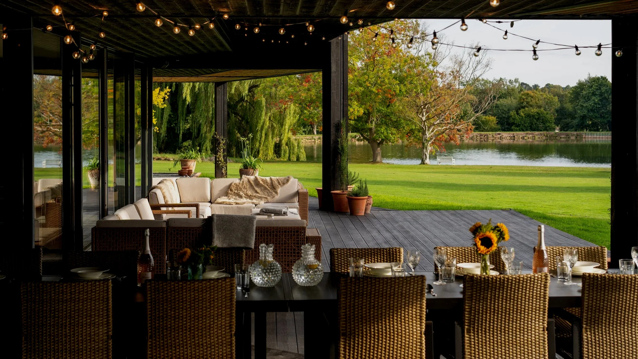 Interior dining area with a large table set for a meal, decorated with sunflowers and bottles, overlooking an outdoor patio and a lush green landscape with trees and a lake.