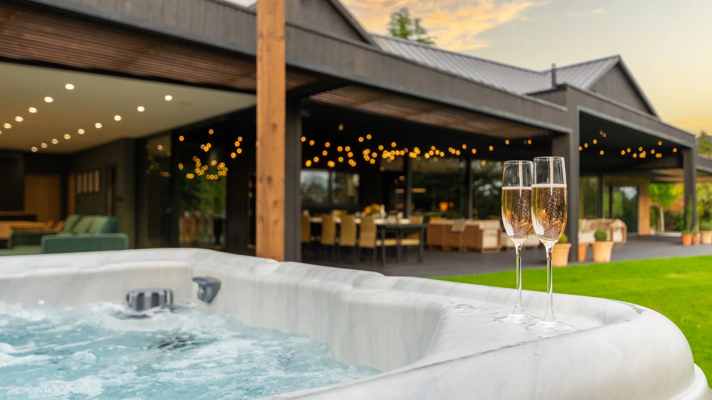 Two glasses of champagne on the edge of a hot tub, with a house and outdoor patio area decorated with string lights in the background at sunset.