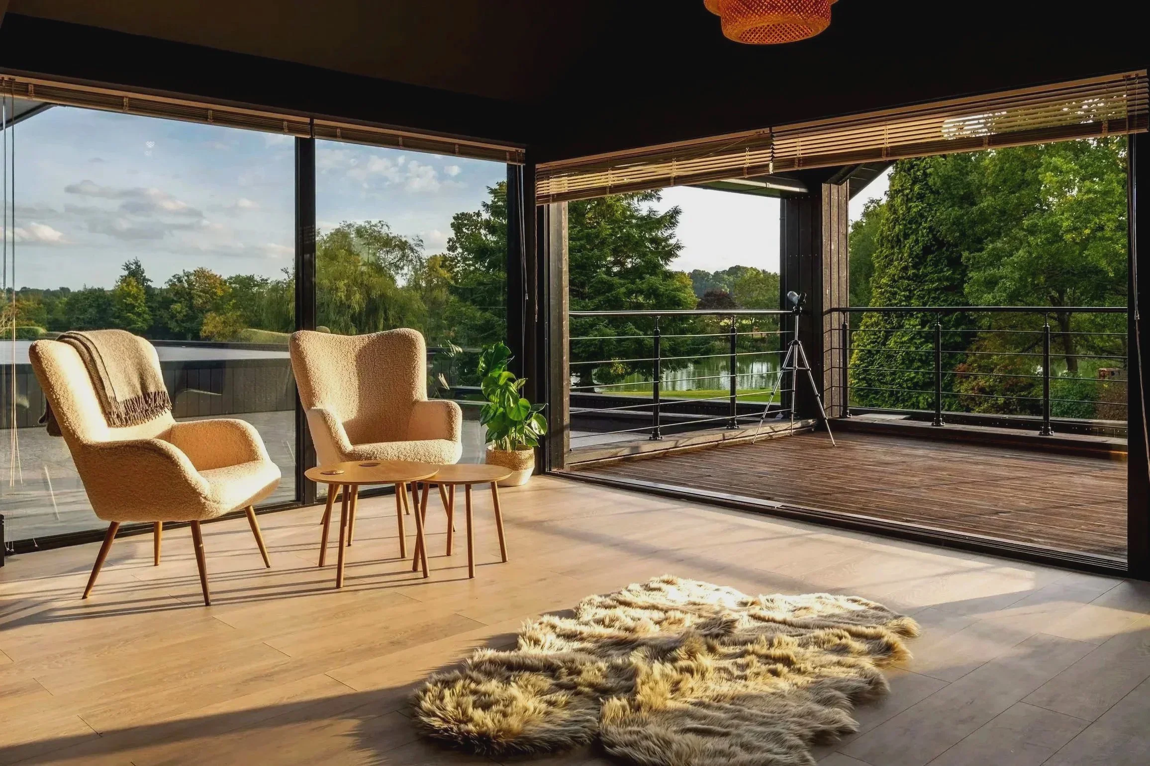 Living room with large floor-to-ceiling windows showing a view of a river and trees, two beige armchairs with brown blankets, a small wooden table between them, a green potted plant, a fur rug on wooden floor, and a telescope on the balcony outside.