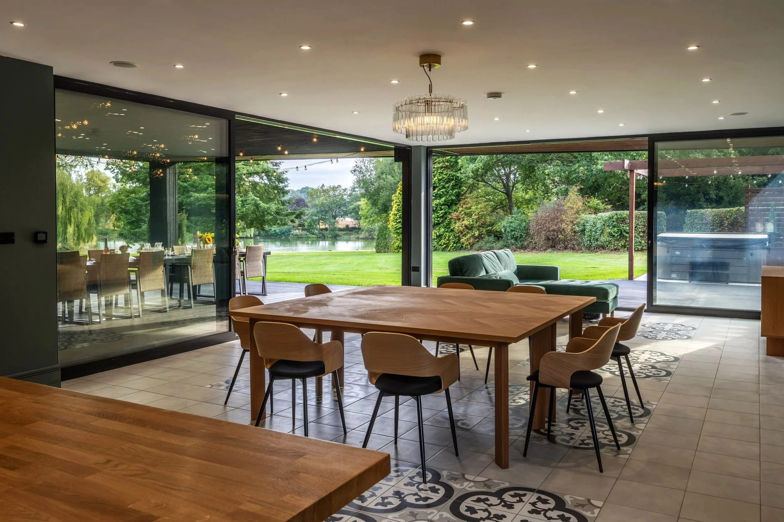 Interior view of a modern home with large glass sliding doors opening to a green outdoor yard with trees and a lake. The room features a wooden dining table with chairs, patterned floor tiles, a sofa, and a chandelier hanging from the ceiling.