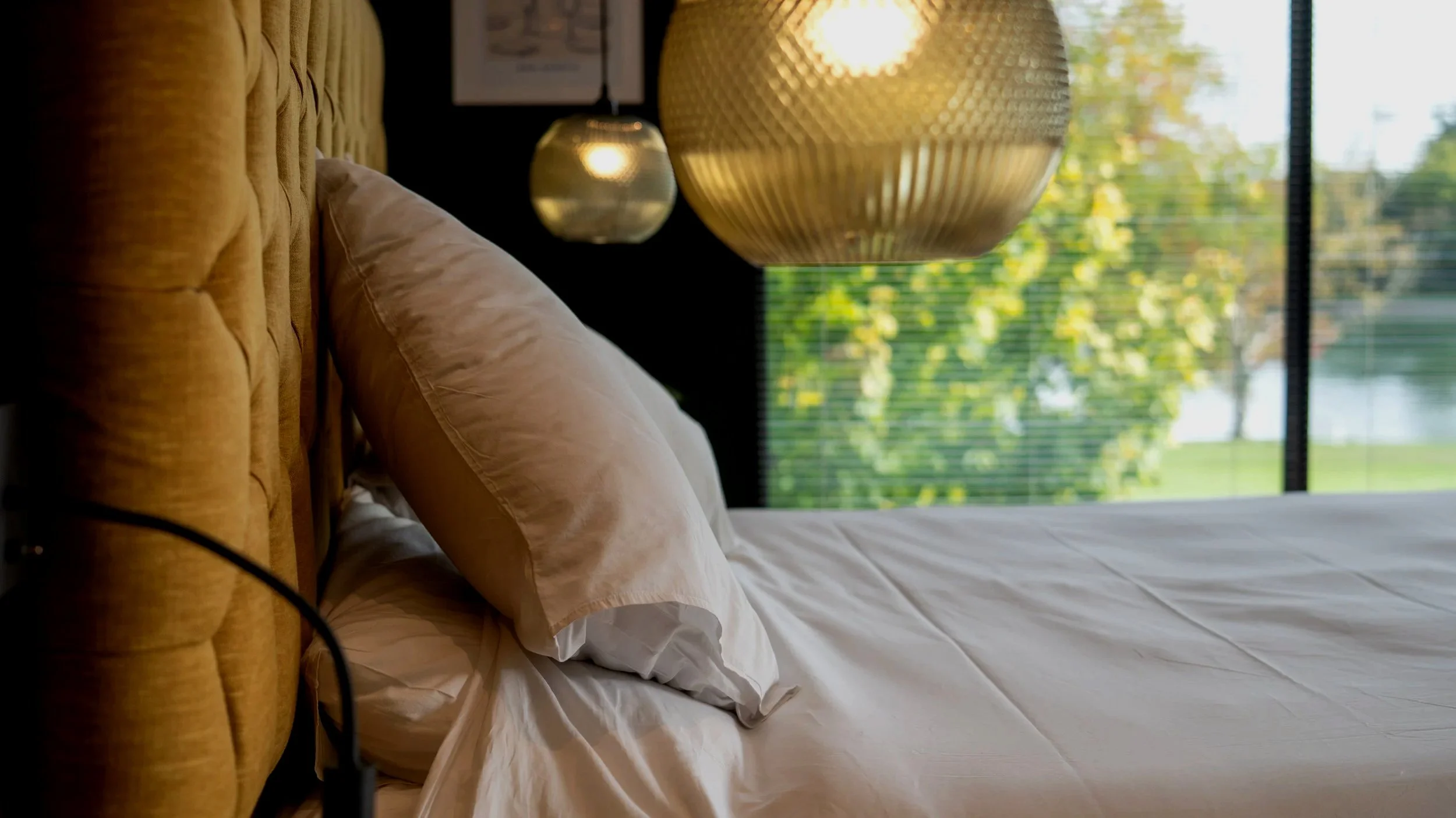 A bedroom with a mustard-colored tufted headboard, white pillows, a white sheet, two hanging pendant lights, and a large window with blinds showing green trees outside.