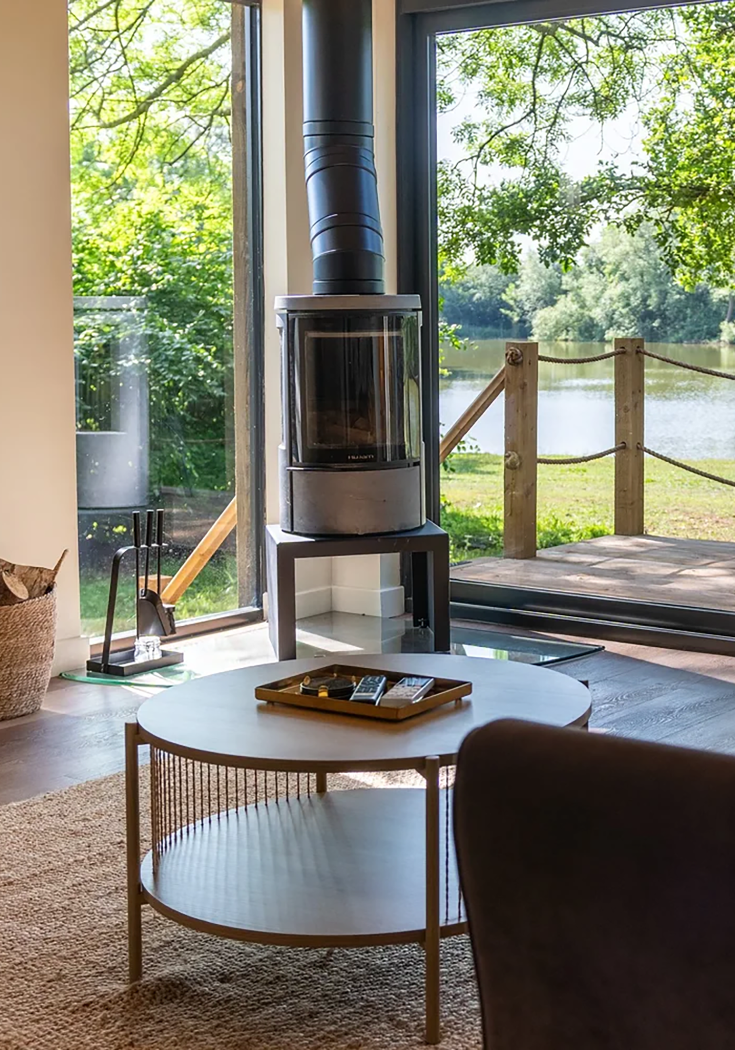 Bright living room with a wood stove near large glass windows overlooking a lake and trees.