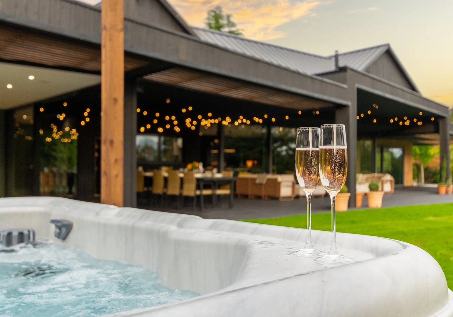 Two glasses of champagne on the edge of an outdoor hot tub, with a house and patio in the background during sunset.