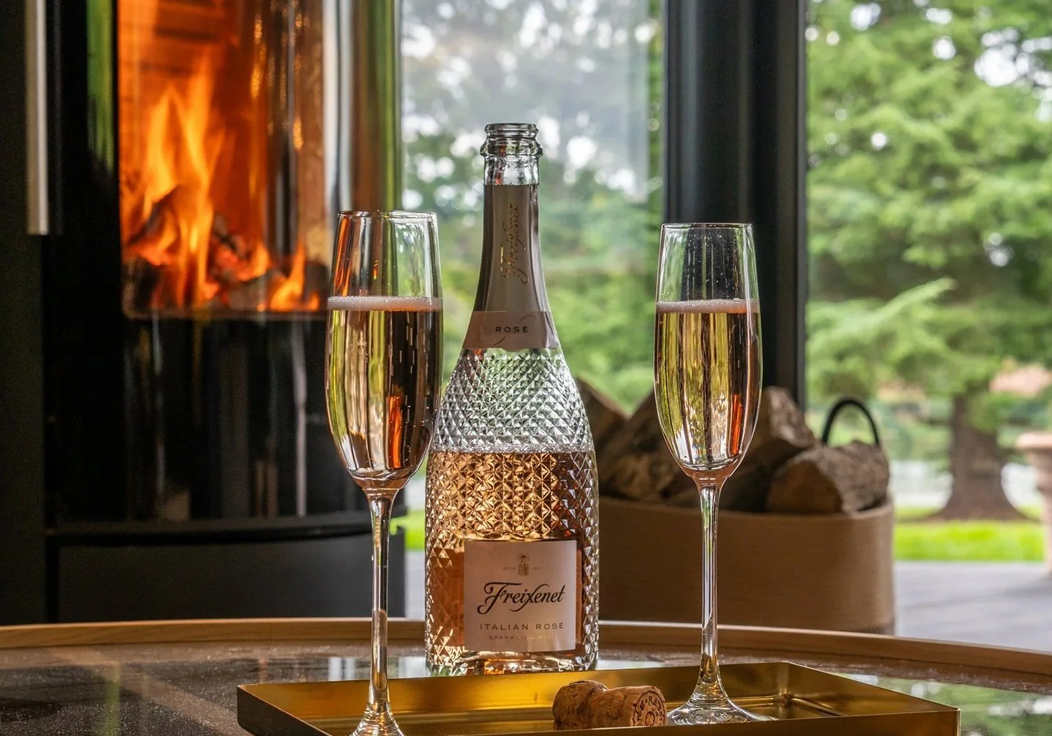 A bottle of Freixenet Italian Rosé sparkling wine with two glasses filled with Rosé on a table. Fireplace and a window with green trees in the background.