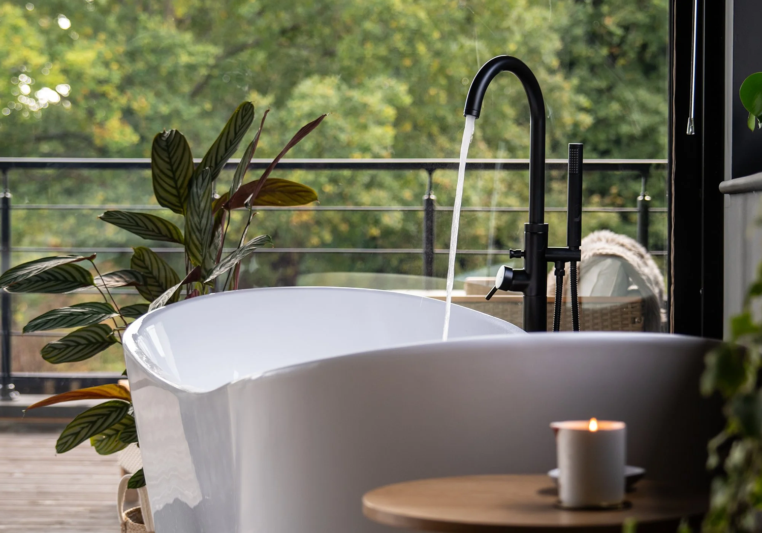 A modern outdoor bathtub placed on a wooden deck, with a black faucet and water running, surrounded by green plants and a lit candle on a small wooden table, overlooking a lush, green forested landscape.