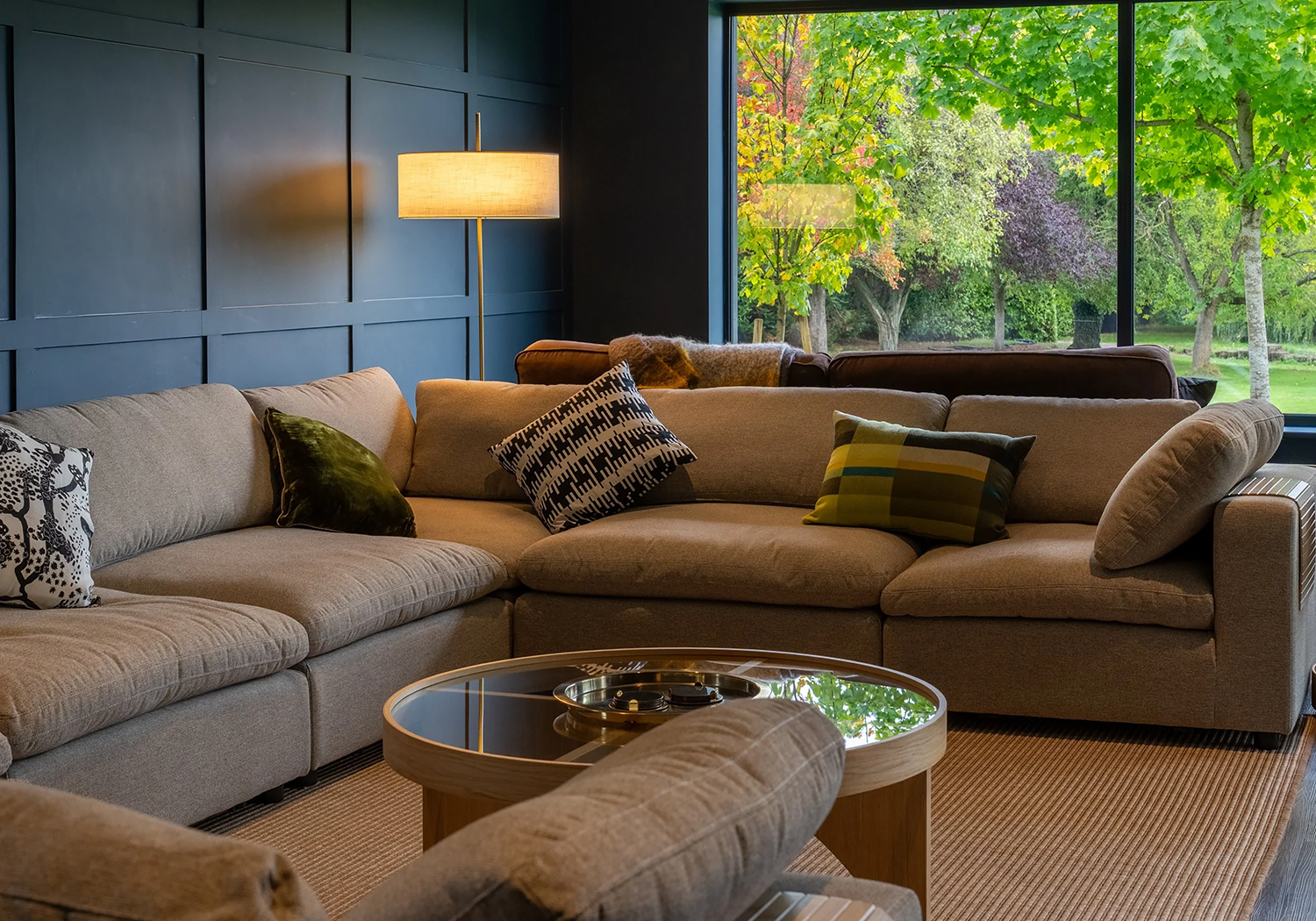 Living room with beige sectional sofa, decorative pillows, a wooden coffee table with a glass top, a floor lamp, and large windows showing a lush outdoor scene with green and autumn-colored trees.