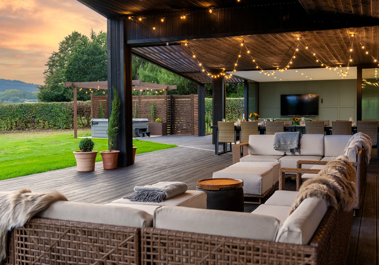 Outdoor patio with string lights, wicker furniture, a beige sectional sofa with blankets, a dining table with chairs, potted plants, and a view of a grassy yard and trees at sunset.
