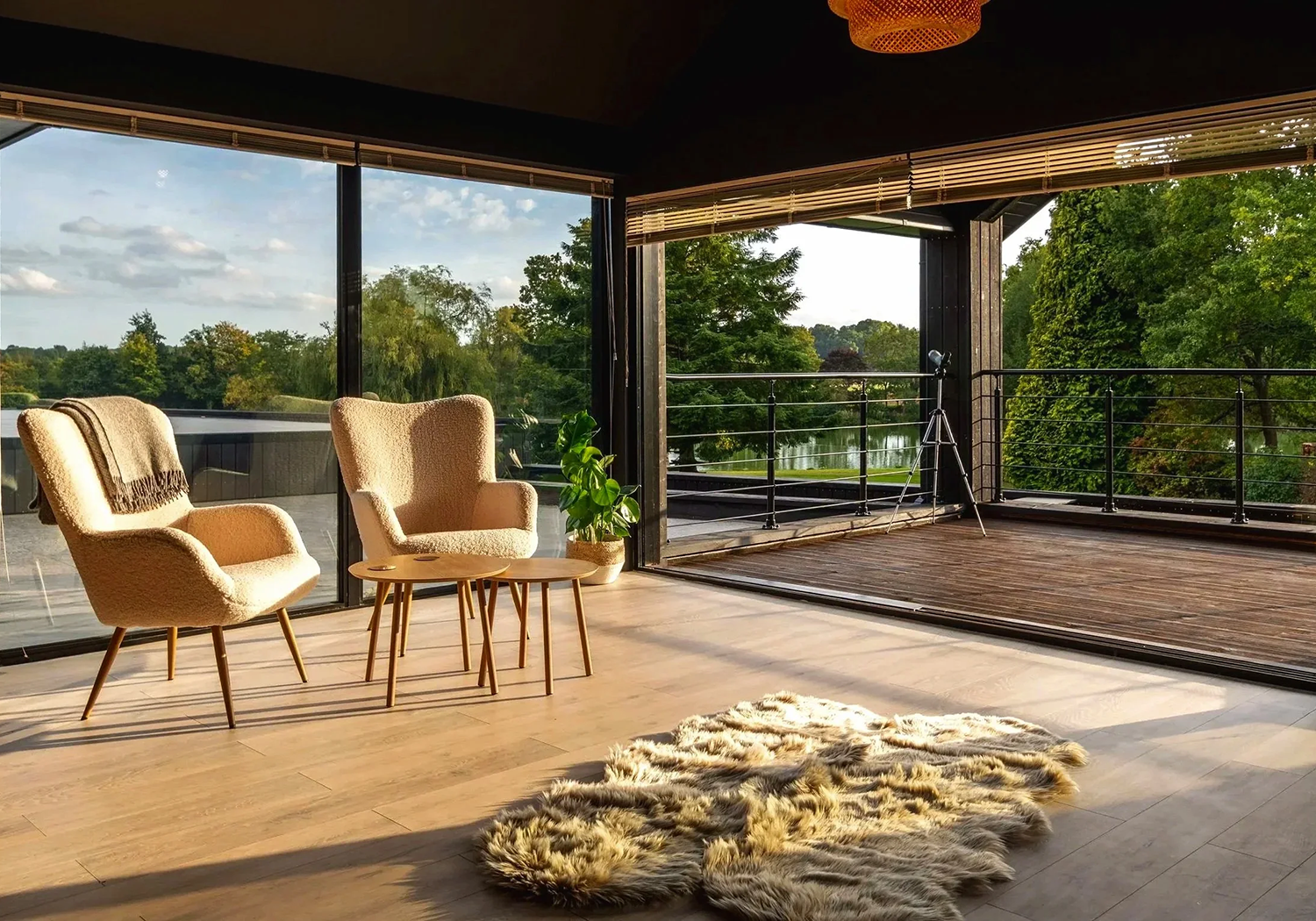 Living room with large glass windows showing a view of trees, a lake, and a partly cloudy sky. There are two beige armchairs with wooden legs, a small wooden side table, a potted plant, and a soft furry rug on the light-colored wooden floor. A telesc