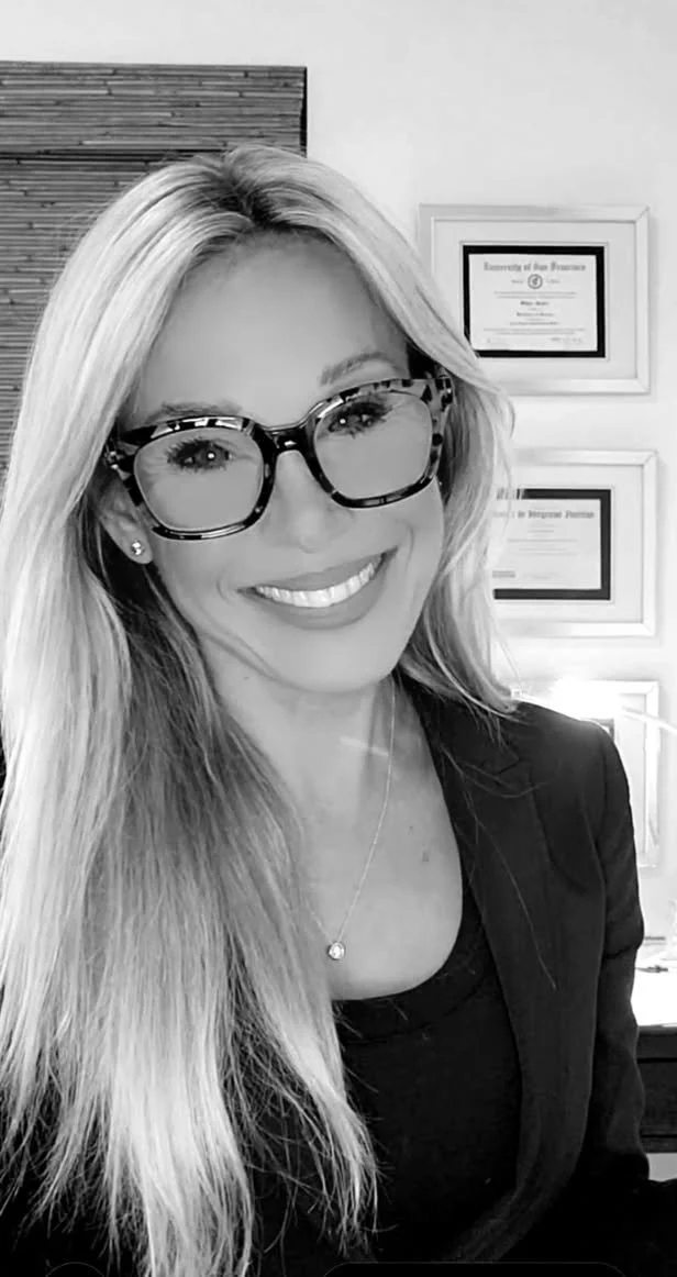 A smiling woman with long blonde hair and large tortoiseshell glasses, wearing a dark blazer and a necklace with a small pendant, in an office with framed certificates on the wall.