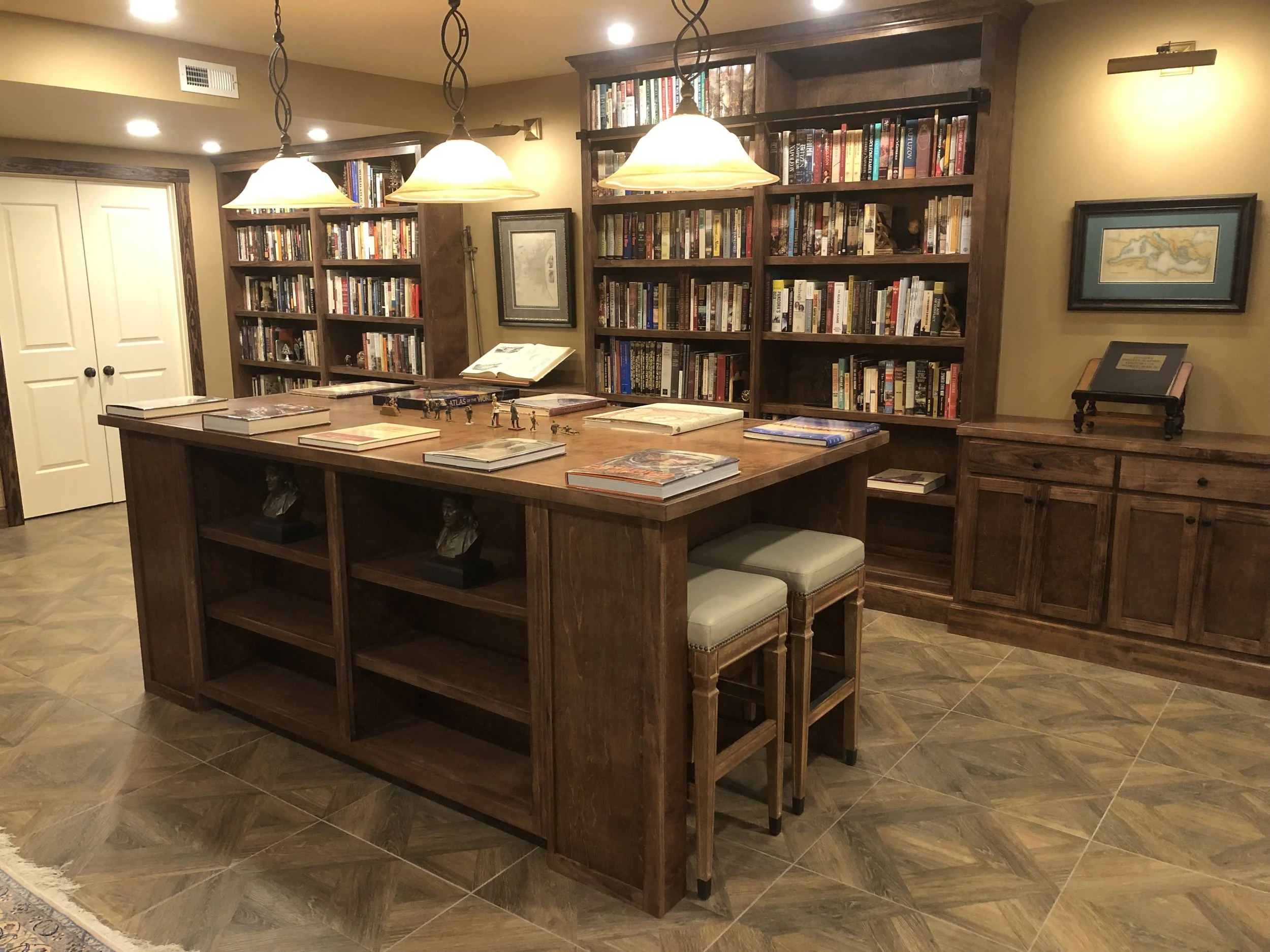 View of a cozy home library or reading room with bookshelves filled with books, a large wooden table, reading chairs, framed artwork on the walls, and pendant lights hanging from the ceiling.