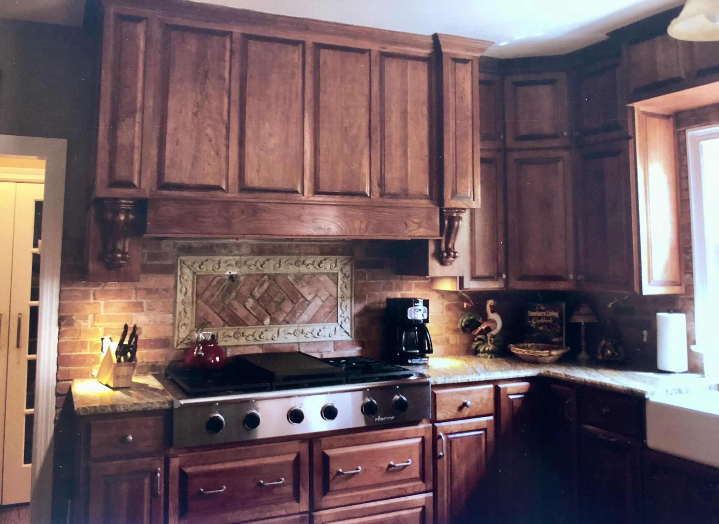 Kitchen with wooden cabinets, brick wall, stove, and countertop. There are decorative items and a window on the right side.