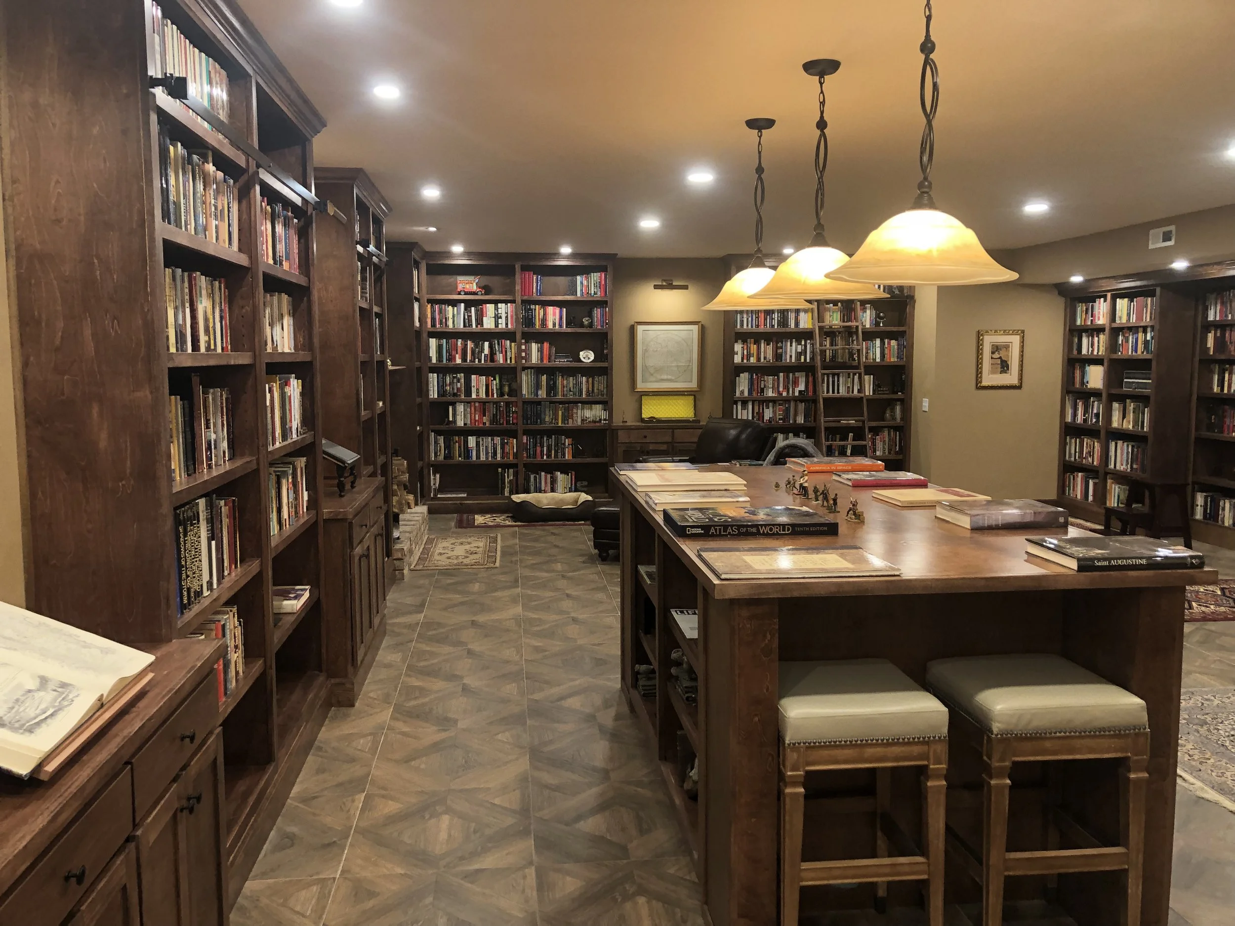 A cozy library with dark wooden bookshelves filled with books, a large wooden table with books and small figurines, and warm lighting from hanging lamps, creating a welcoming atmosphere for reading.