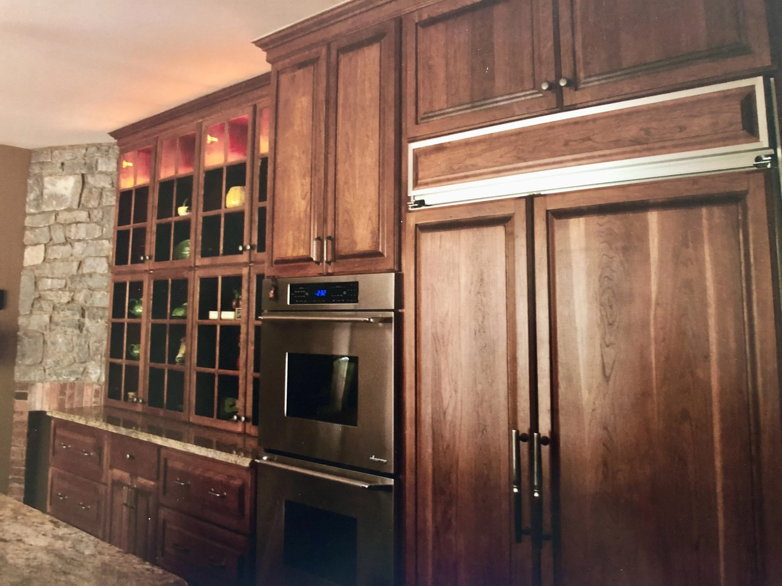 Wooden kitchen cabinets with some open shelf space showing decorative items, a stone wall on the left, and a black stove underneath the cabinets.