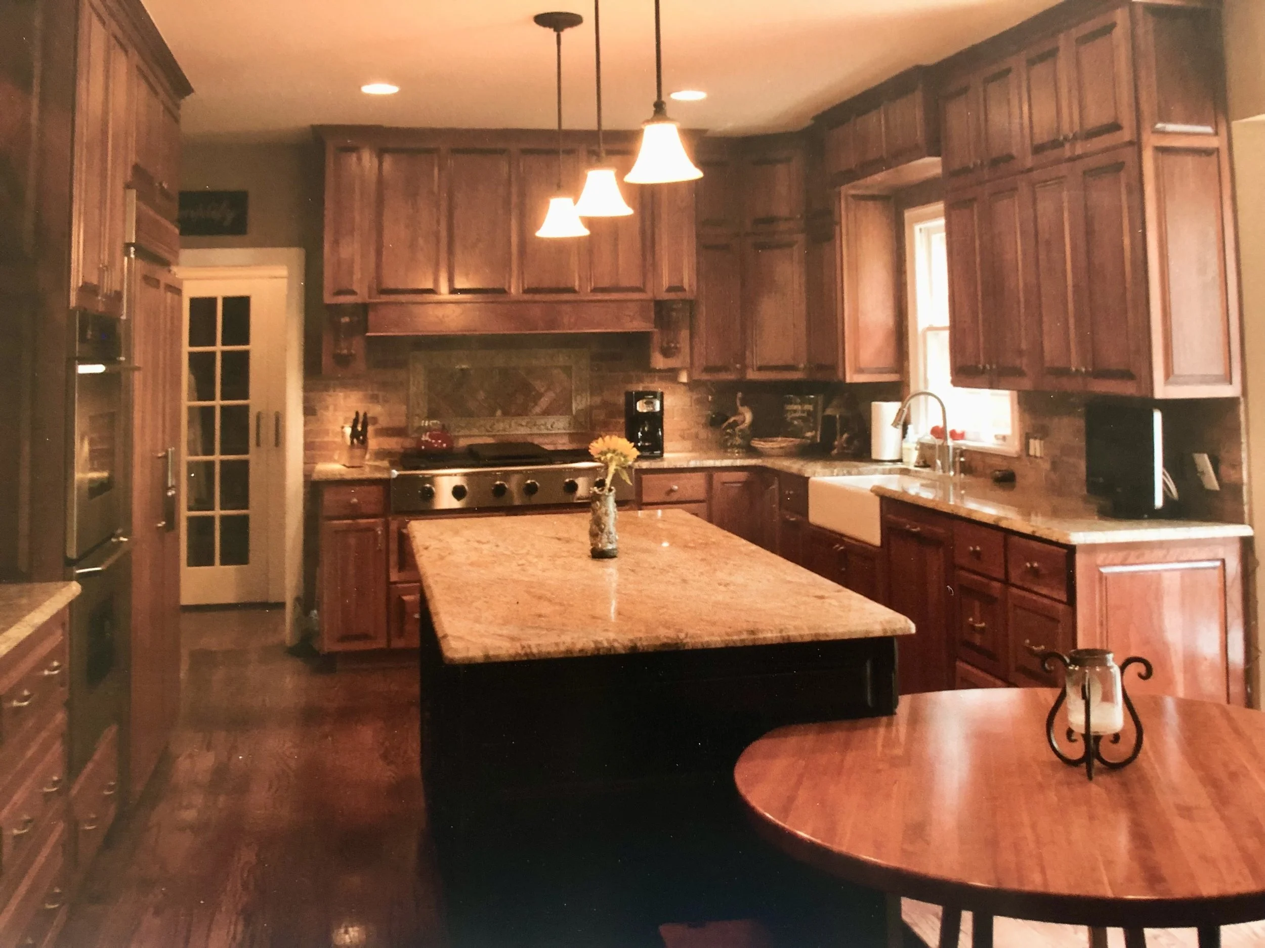 A kitchen with wooden cabinets, a kitchen island with a granite top, and pendant lights hanging from the ceiling. There is a window above the sink, and a brick backsplash over the stove. The space is illuminated by warm lighting.