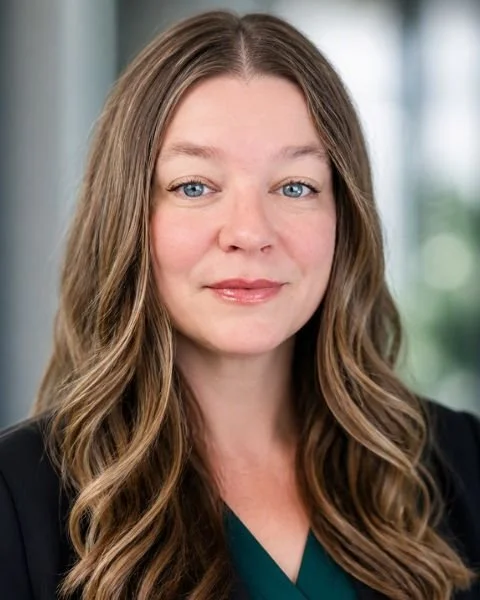 Portrait of a woman with long wavy light brown hair, blue eyes, and fair skin, wearing a dark blazer and a green top, standing indoors with a blurred background.