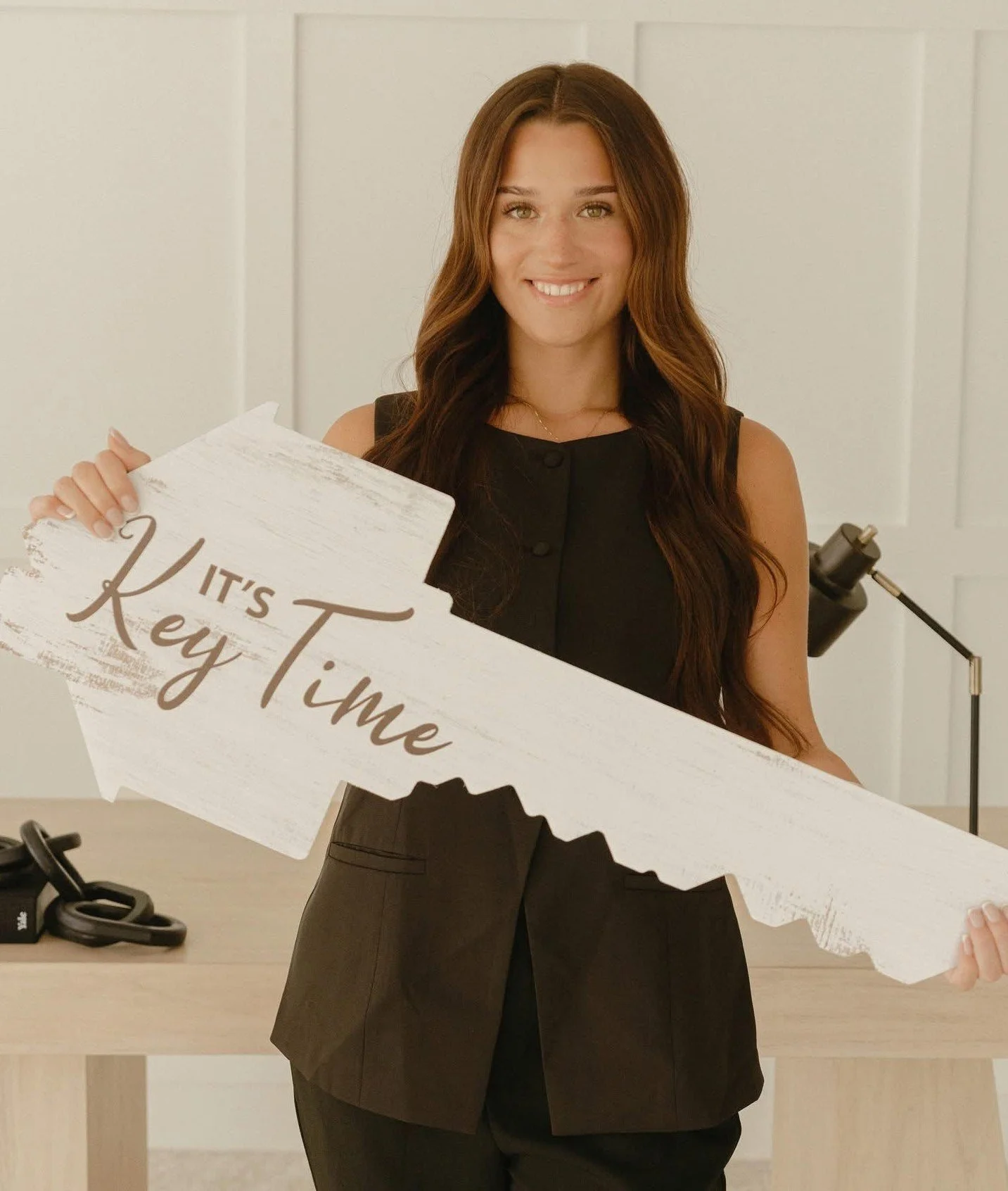 A woman with long, wavy brown hair smiling while holding a large white arrow sign that says "It's Key Time" in brown lettering. She is wearing a sleeveless black top and brown pants in a bright room with a white paneled wall in the background.