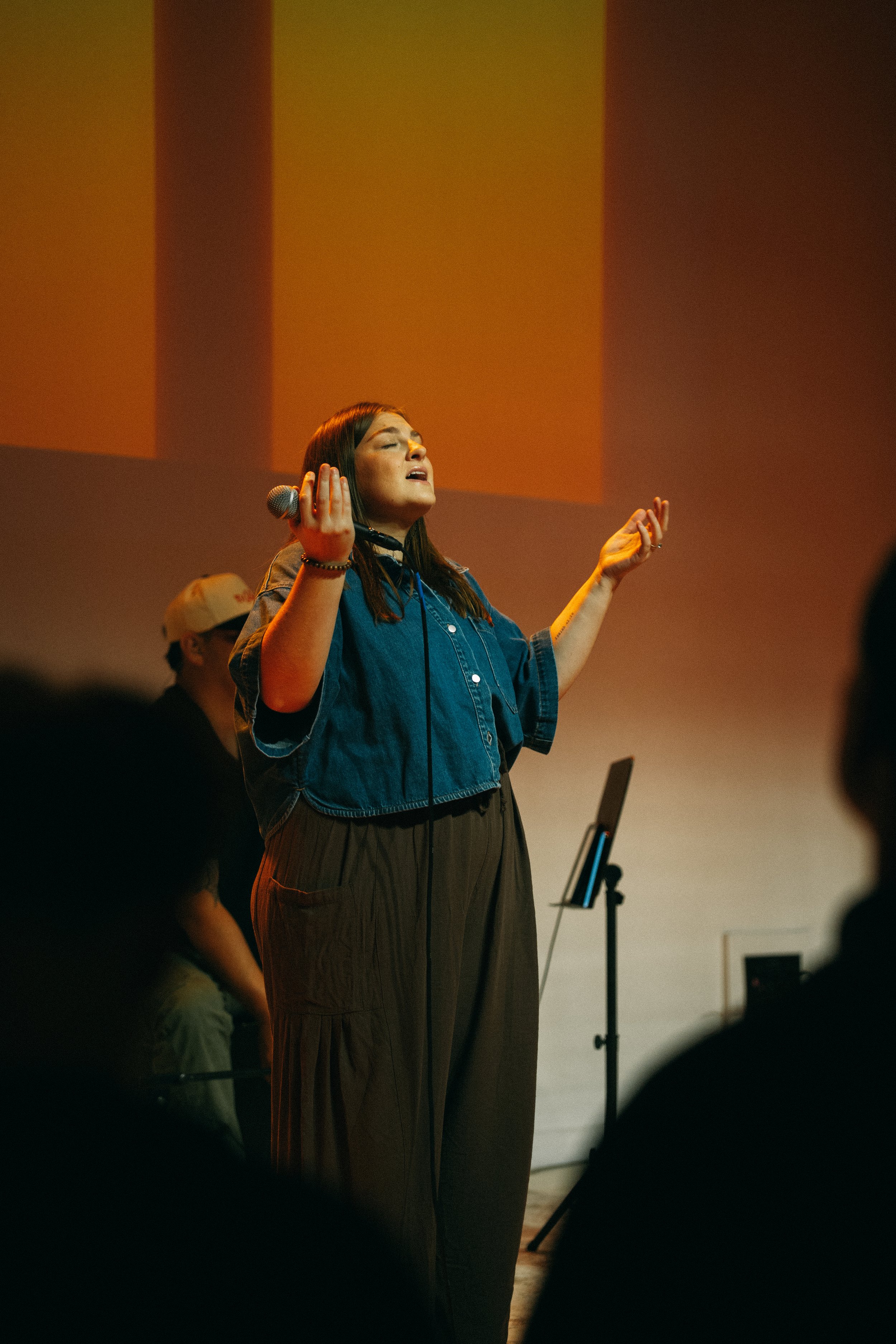 A woman singing into a microphone with her eyes closed, raising her hands, in a dimly lit room with a large screen behind her and a person in a hat and glasses standing nearby.