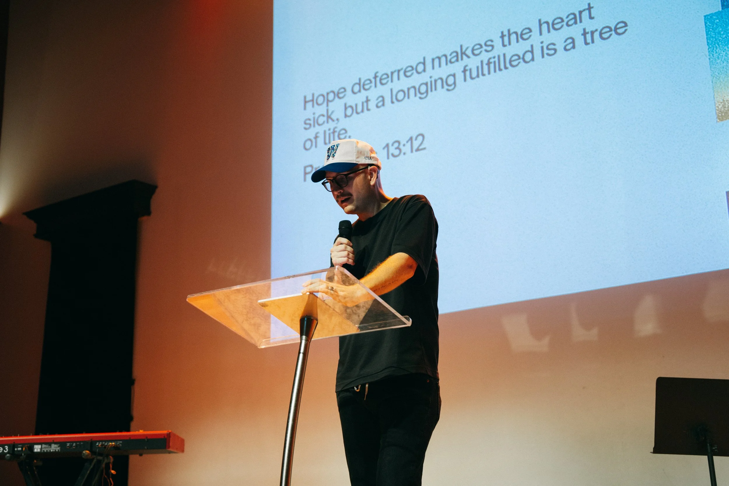 A man wearing glasses and a white cap with colorful patterns is speaking into a microphone at a podium on a stage. Behind him, there is a large screen displaying text with a passage of scripture.
