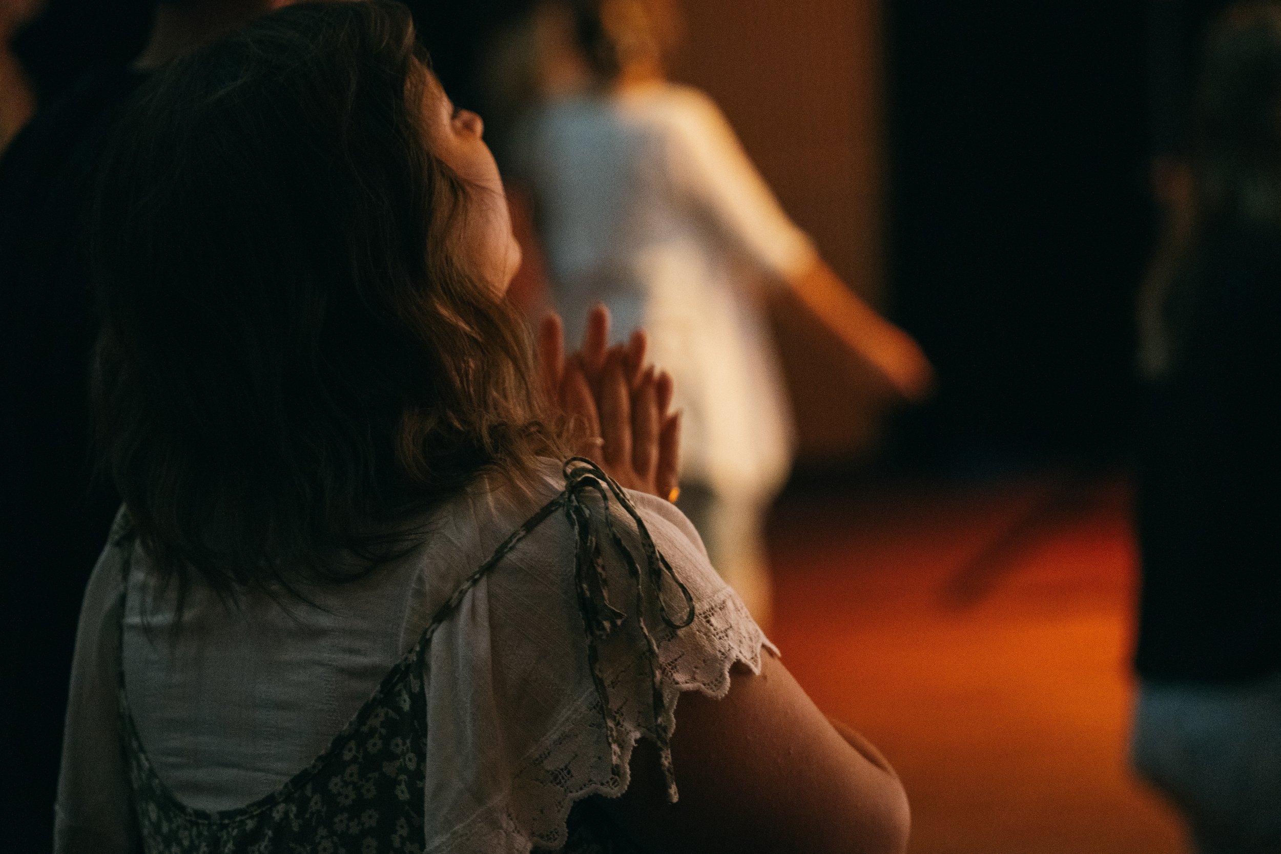 A woman with long curly hair and wearing a lace and patterned dress appears to be in a prayerful posture, with her hands clasped in front of her face. The background is dark with blurred figures, suggesting an indoor setting such as a church or a gathering.