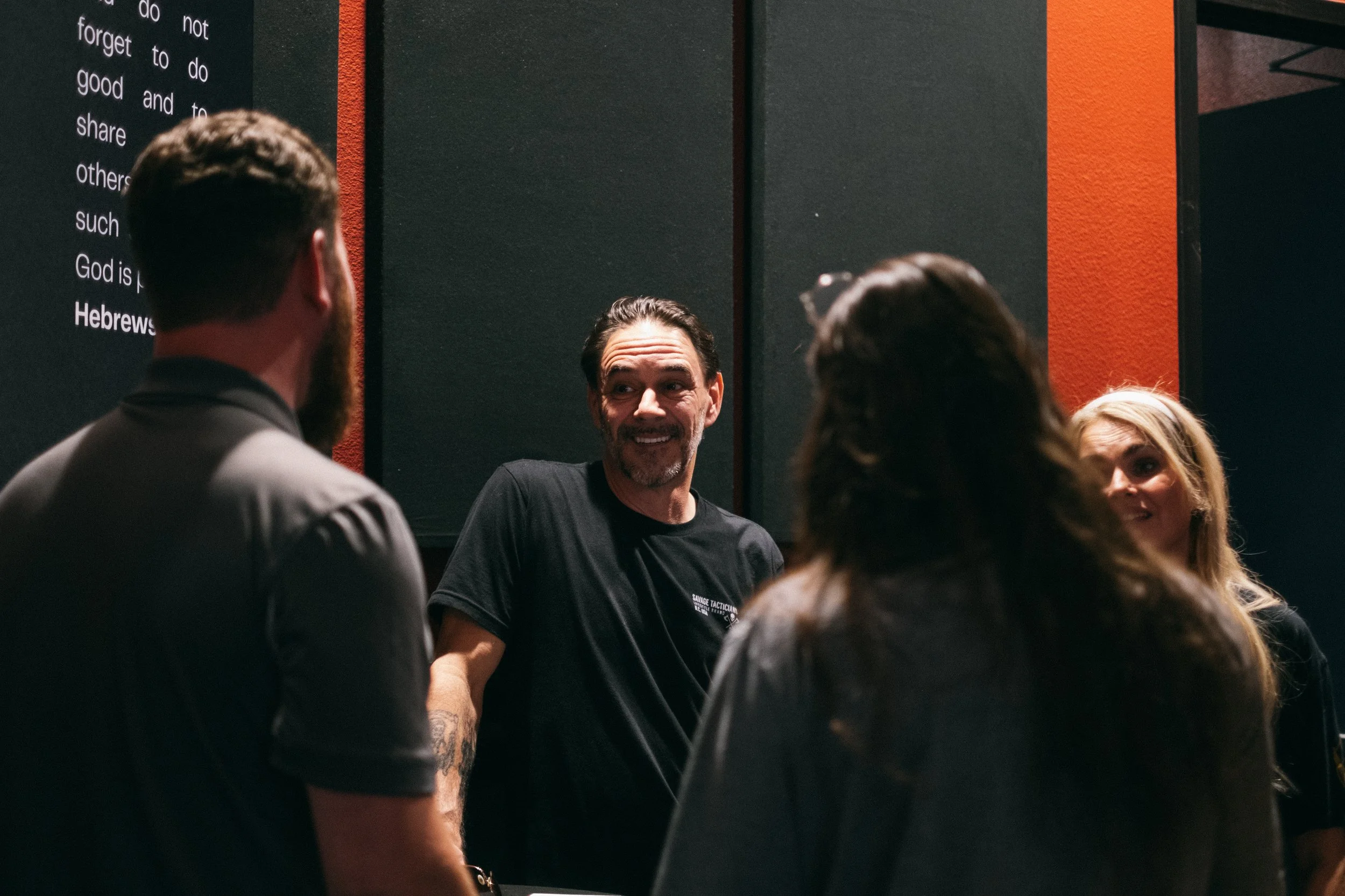 A group of people having a conversation indoors, with a man in a black T-shirt smiling and talking to others, with black and red walls in the background.