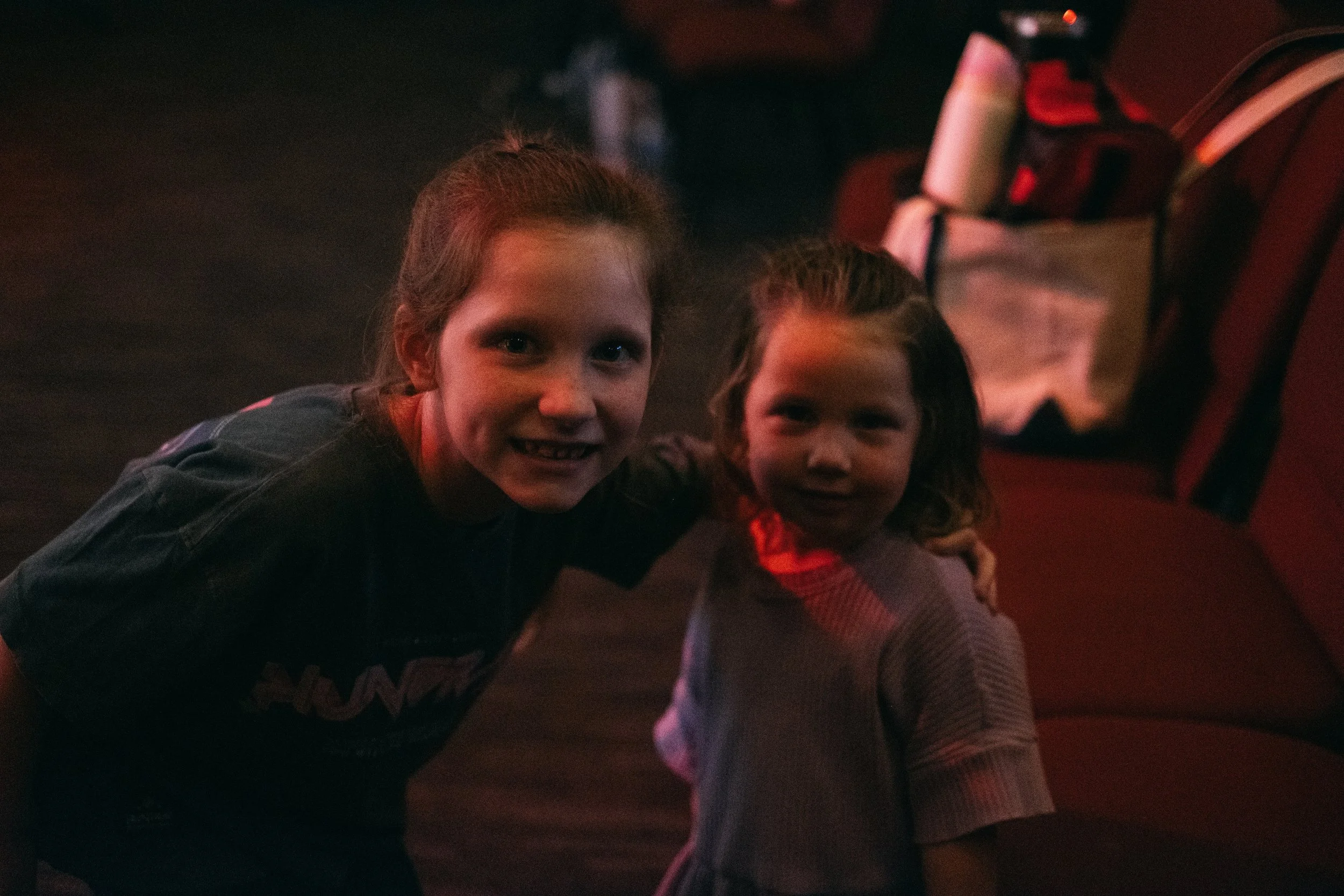 Two young girls with brown hair sitting close together in a dimly lit environment, smiling at the camera.