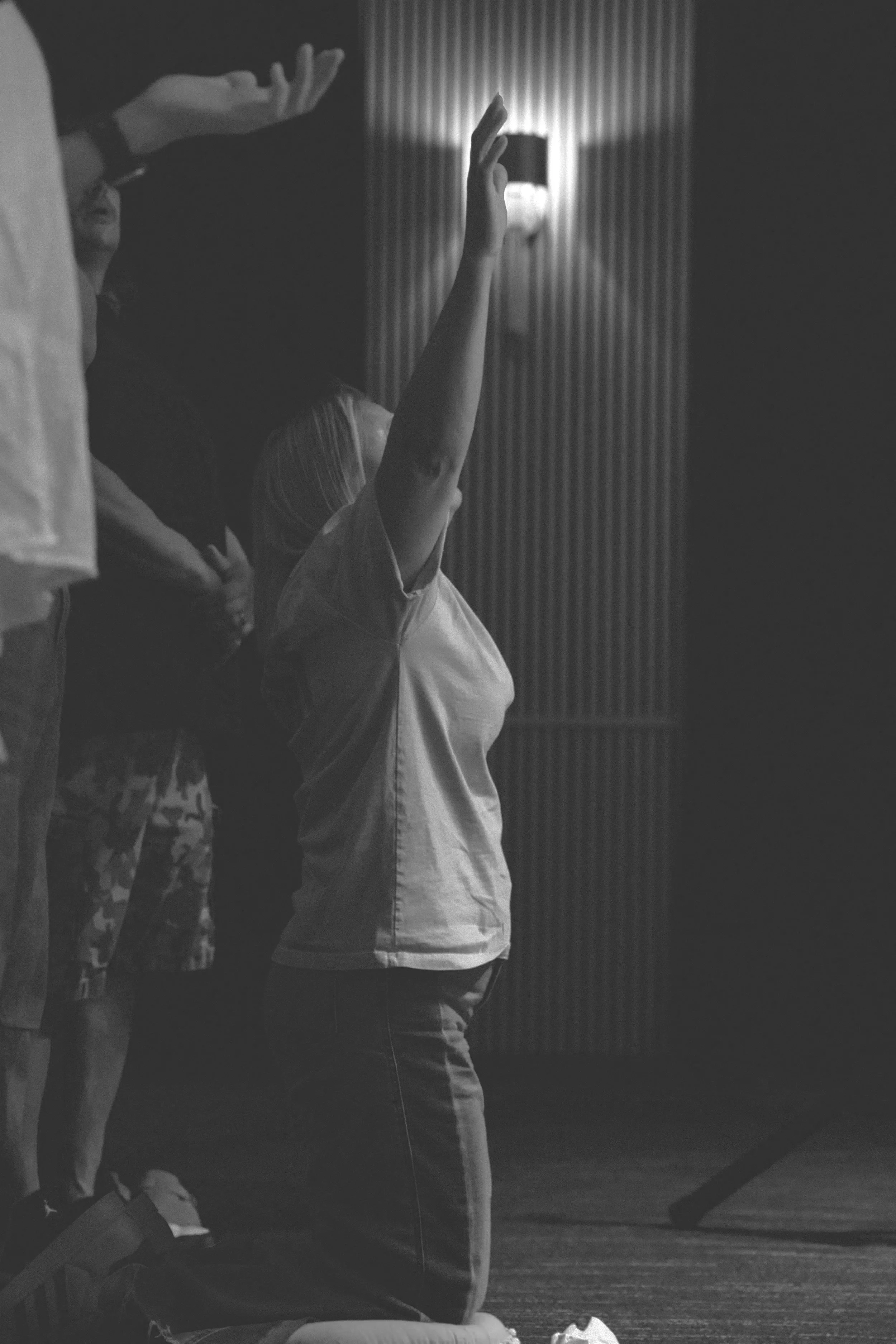 A person on their knees with their hand raised towards the sky, in a dimly lit room.