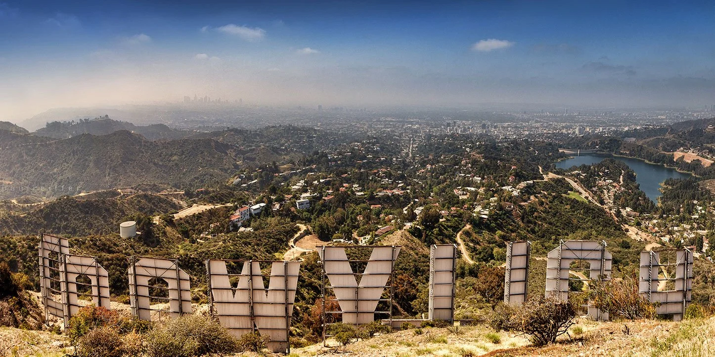 A panoramic view of Los Angeles from the Hollywood Hills, with the Hollywood sign in the foreground and city skyline in the background, under a partly cloudy sky.