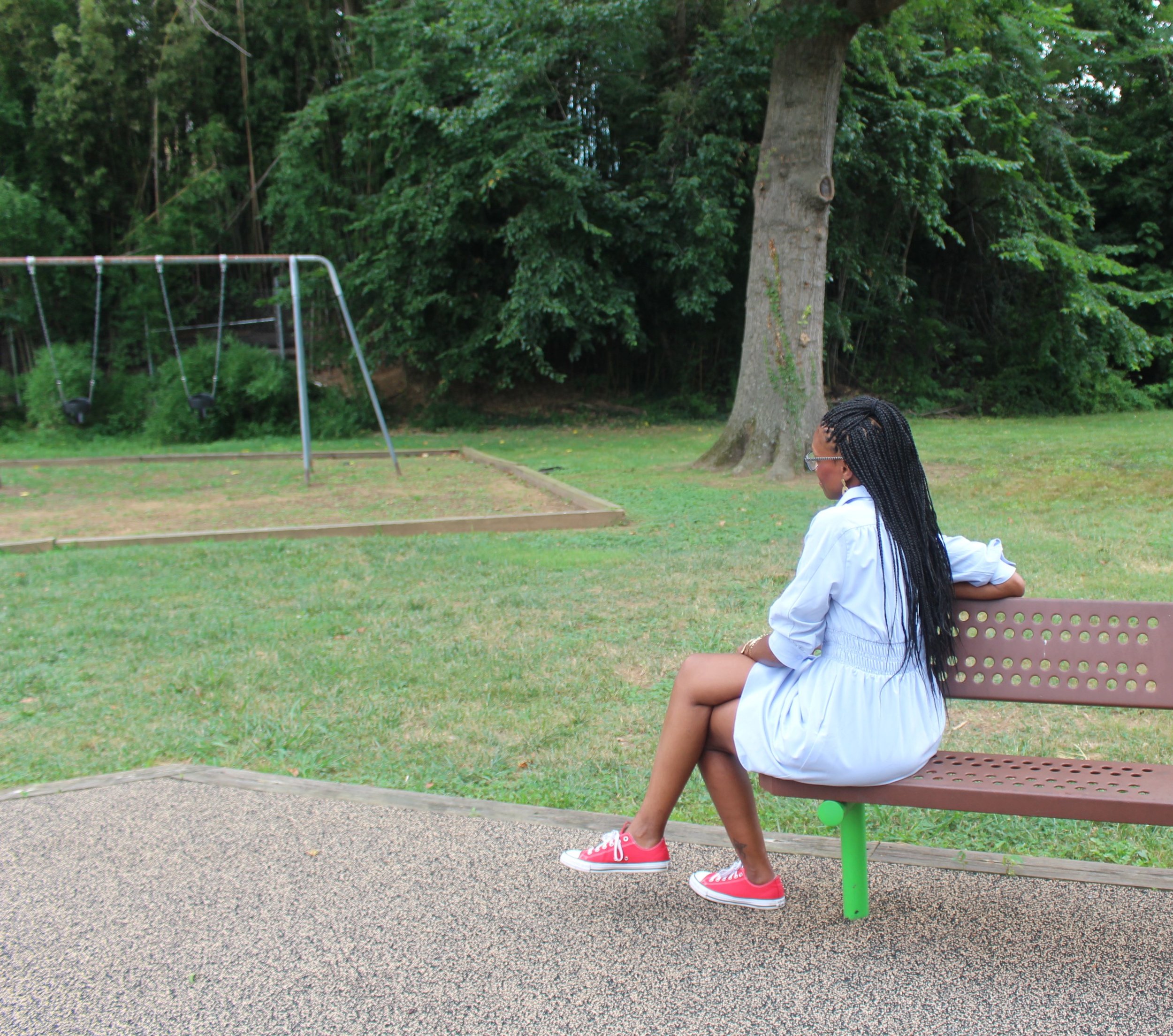 A young girl with long braided hair wearing a white dress and pink shoes sitting on a pink park bench in a park, with playground equipment and trees in the background.