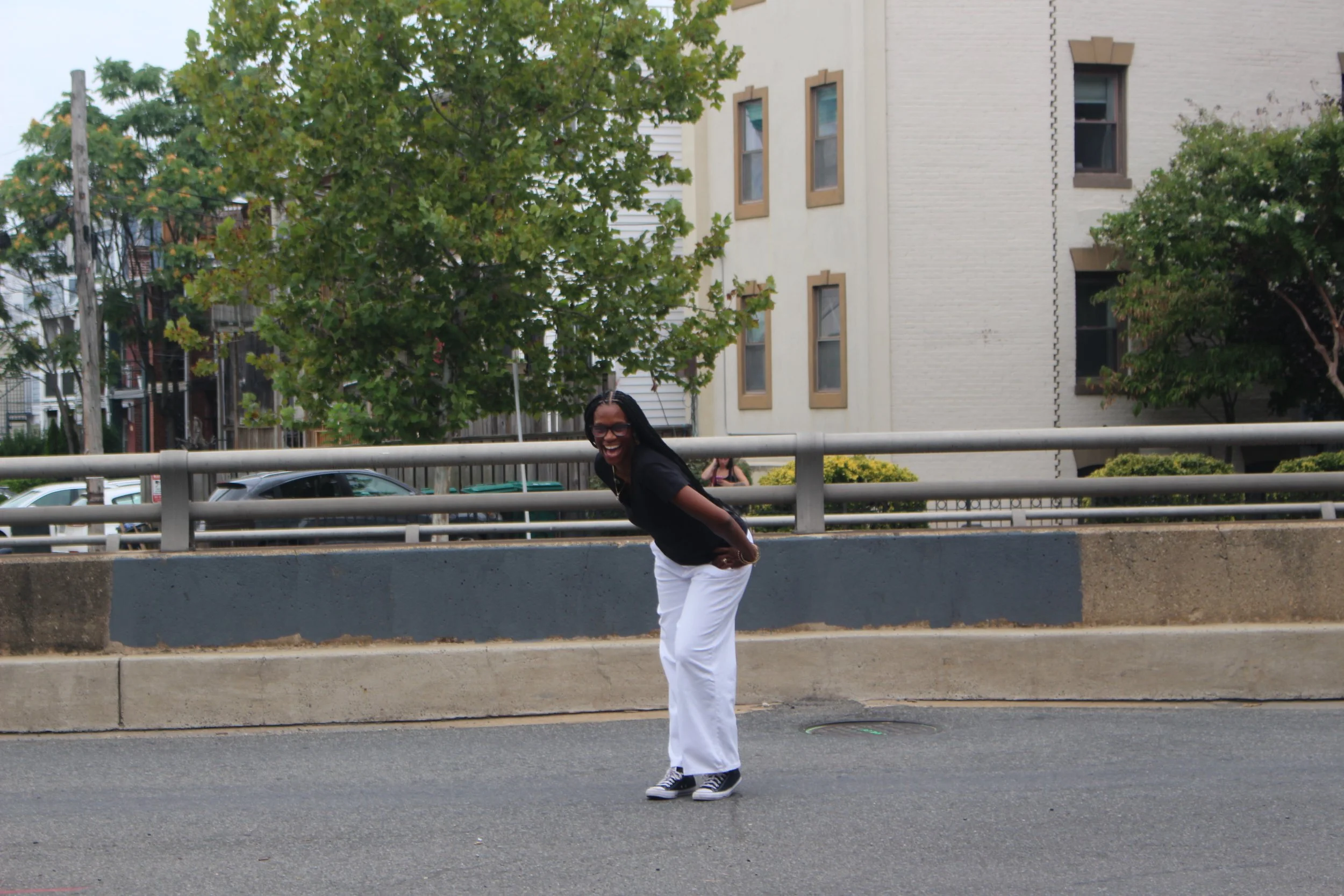 A person with glasses and long hair smiling and leaning forward on a metal railing on a city street, wearing a black top, white pants, and sneakers.