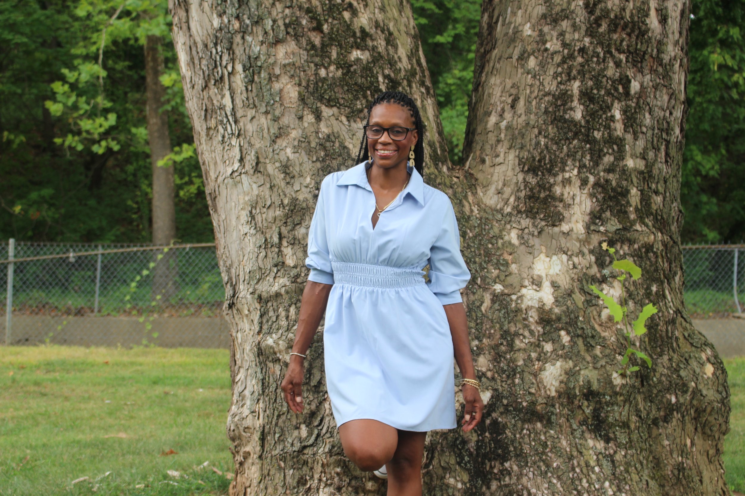 A woman with dark braided hair and glasses smiles while standing in front of a large tree in a park, wearing a light blue dress and earrings.