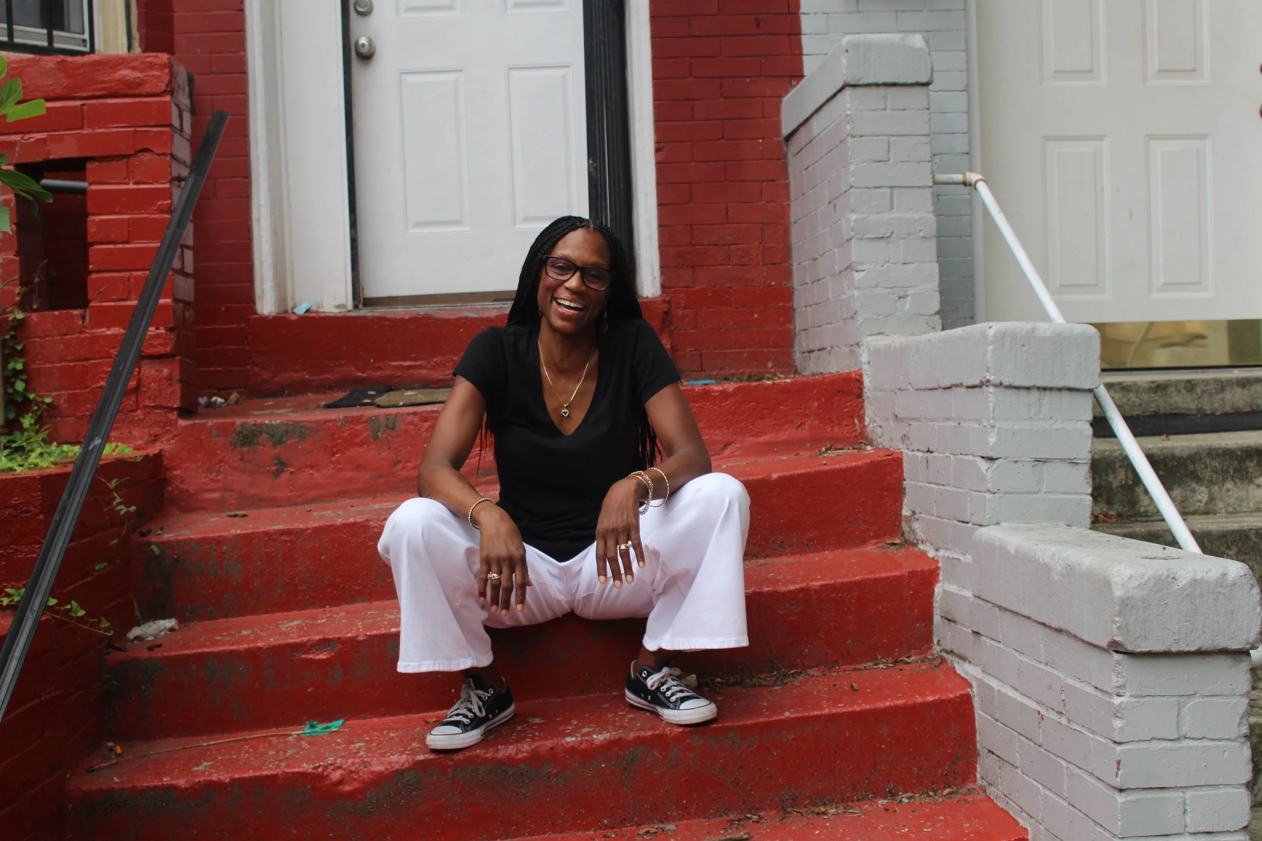 A woman with black braids, glasses, wearing a black t-shirt, white pants, and black sneakers, sitting on red-painted concrete steps in front of a brick building with a white door, smiling and looking at the camera.