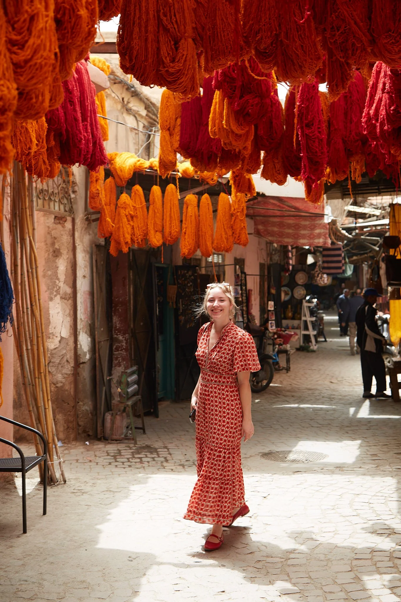 A woman in a red patterned dress and red shoes standing on a cobblestone street in what appears to be a market or shopping area, with hanging yarn or textile strands above her.