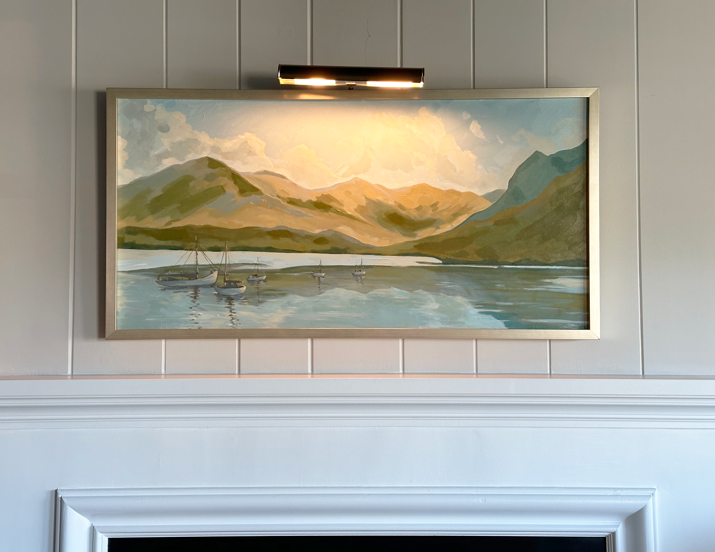 A framed landscape painting of mountains, clouds, and boats on water hangs on a gray wall with an overhead light fixture illuminating it.