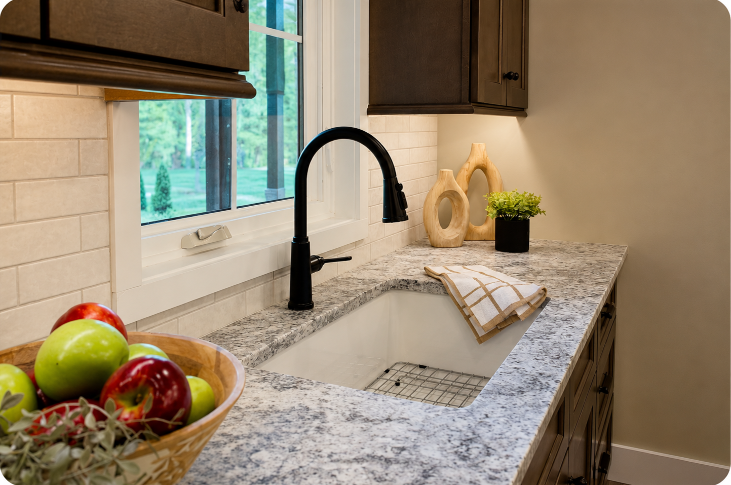Kitchen countertop with a sink, black faucet, a basket of apples, decorative wooden vases, a potted plant, a window showing greenery outside, and beige tile backsplash.