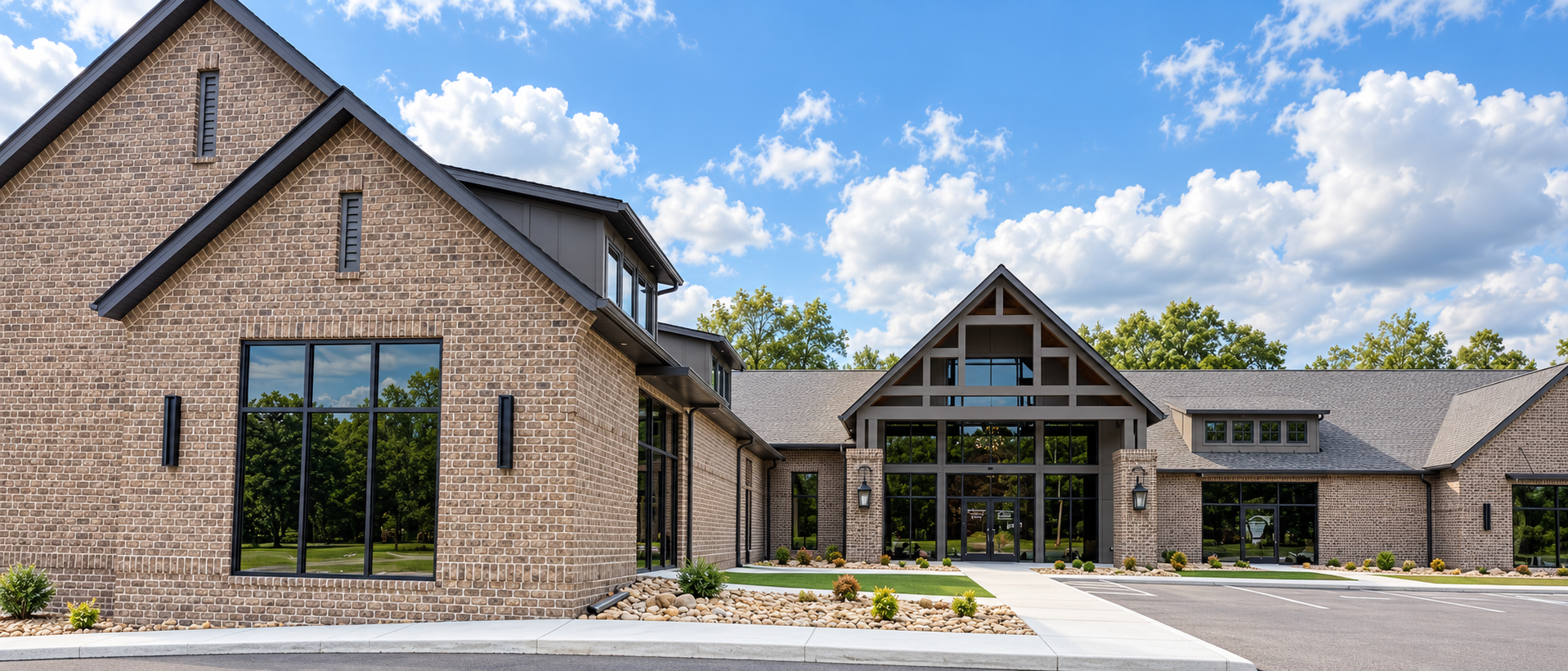 A modern brick building with large glass windows, surrounded by a landscaped area with small bushes and rocks, under a partly cloudy sky.