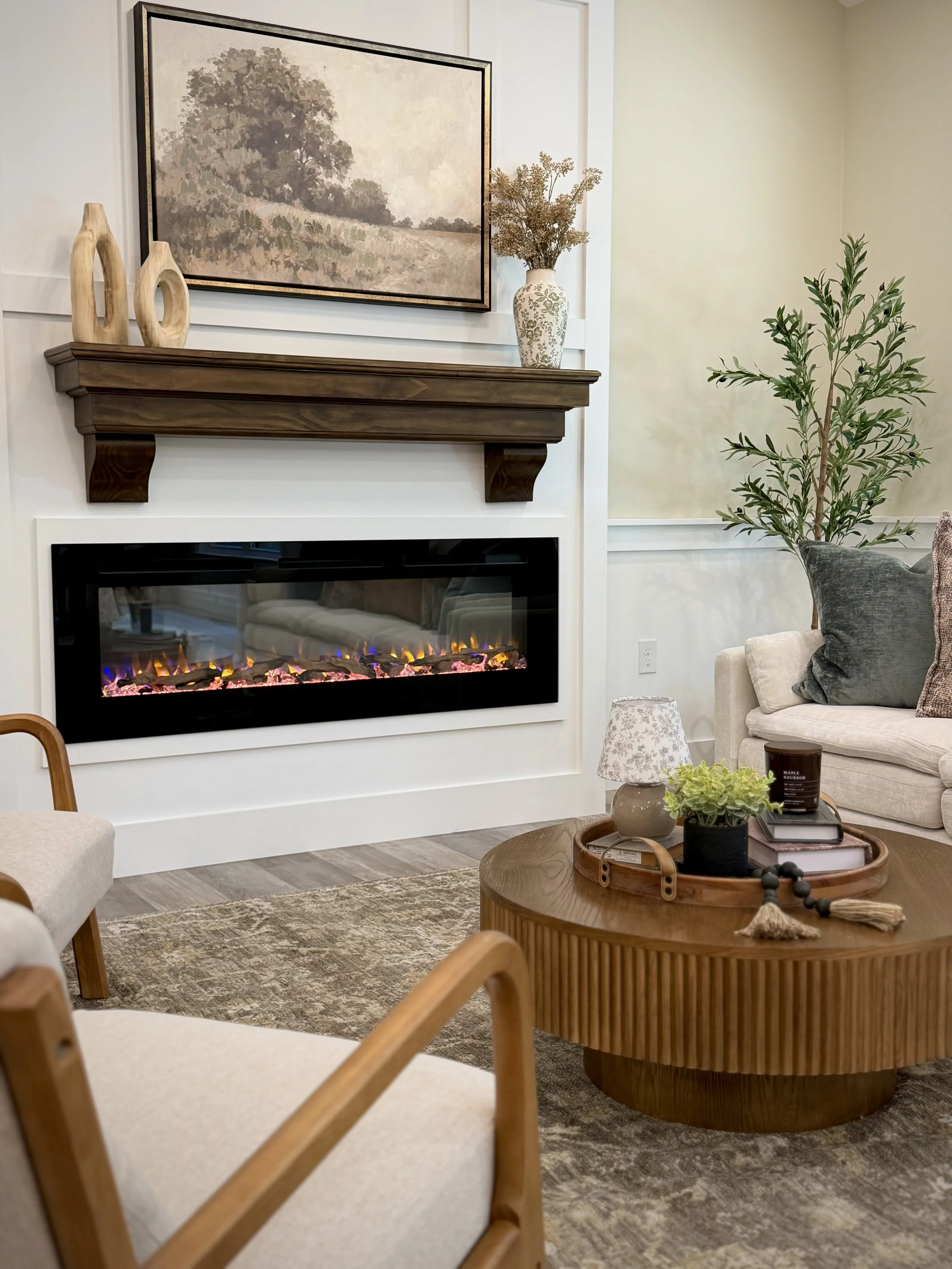 Living room with a wall-mounted electric fireplace, wooden mantel decor with vases, a large landscape painting, a beige sofa with grey and patterned pillows, a wooden coffee table with decorative items including a plant in a black pot, books, a candle, and a small ceramic lamp, and a tall plant in the corner.