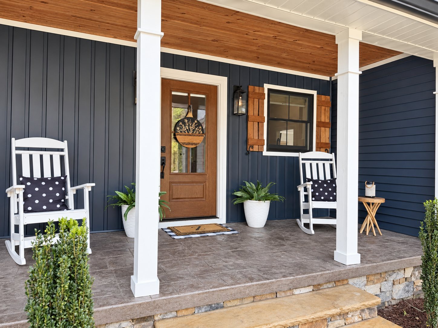Front porch of a house with a wooden door, black siding, and white trim. Two white rocking chairs with polka dot pillows, a small side table, potted plants, and a welcome mat are on the porch.