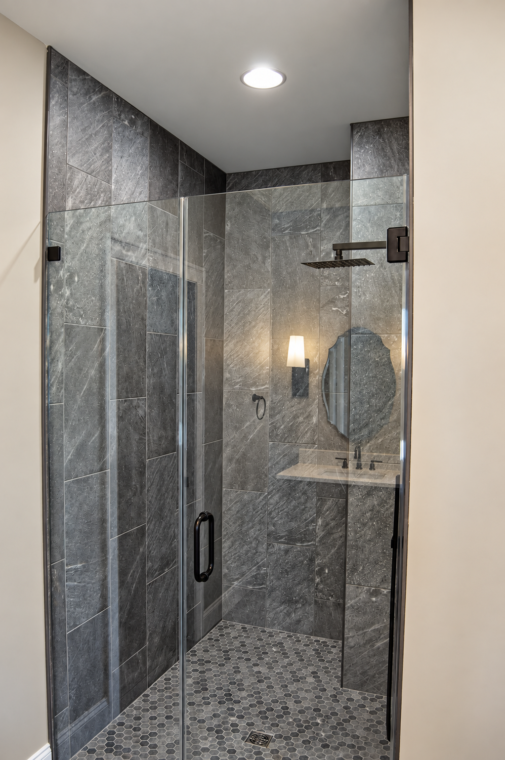 Modern gray tiled walk-in shower with glass door, wall-mounted showerhead, and small hexagonal floor tiles.