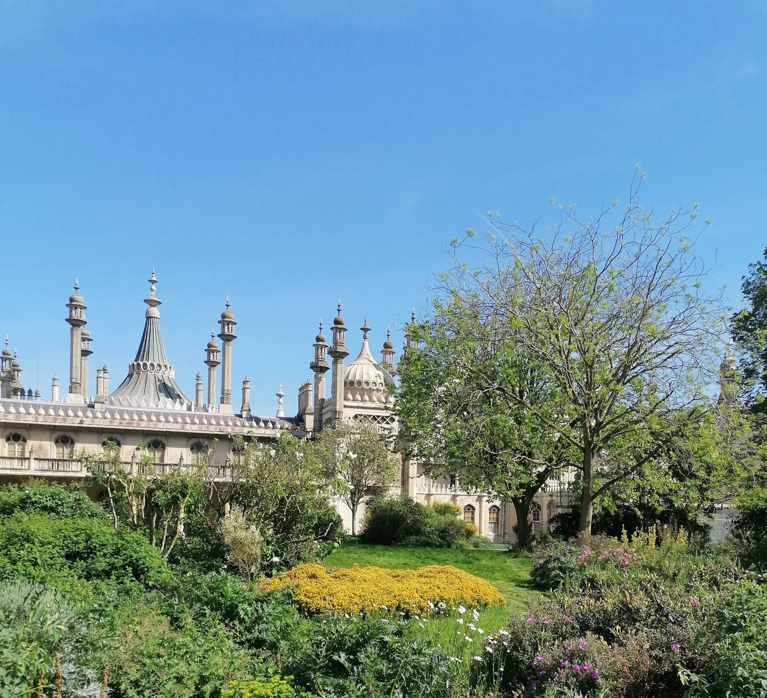 A castle with multiple spires and domes, surrounded by lush green garden and trees under a clear blue sky.