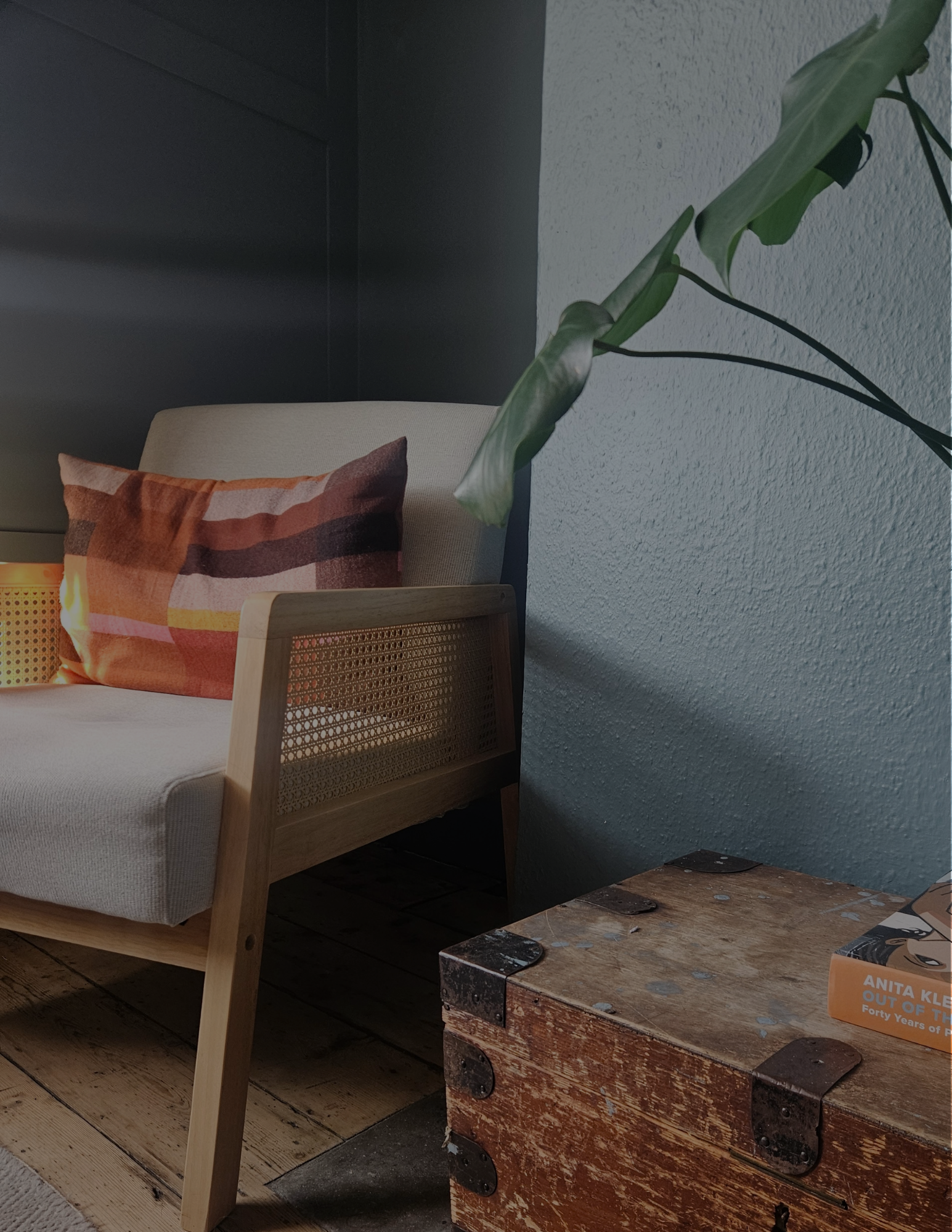 Close-up photo of a beige armchair with a colorful striped pillow, a small lamp on a side table, and a large green plant with broad leaves. A rustic wooden chest and a small box are visible in the foreground.
