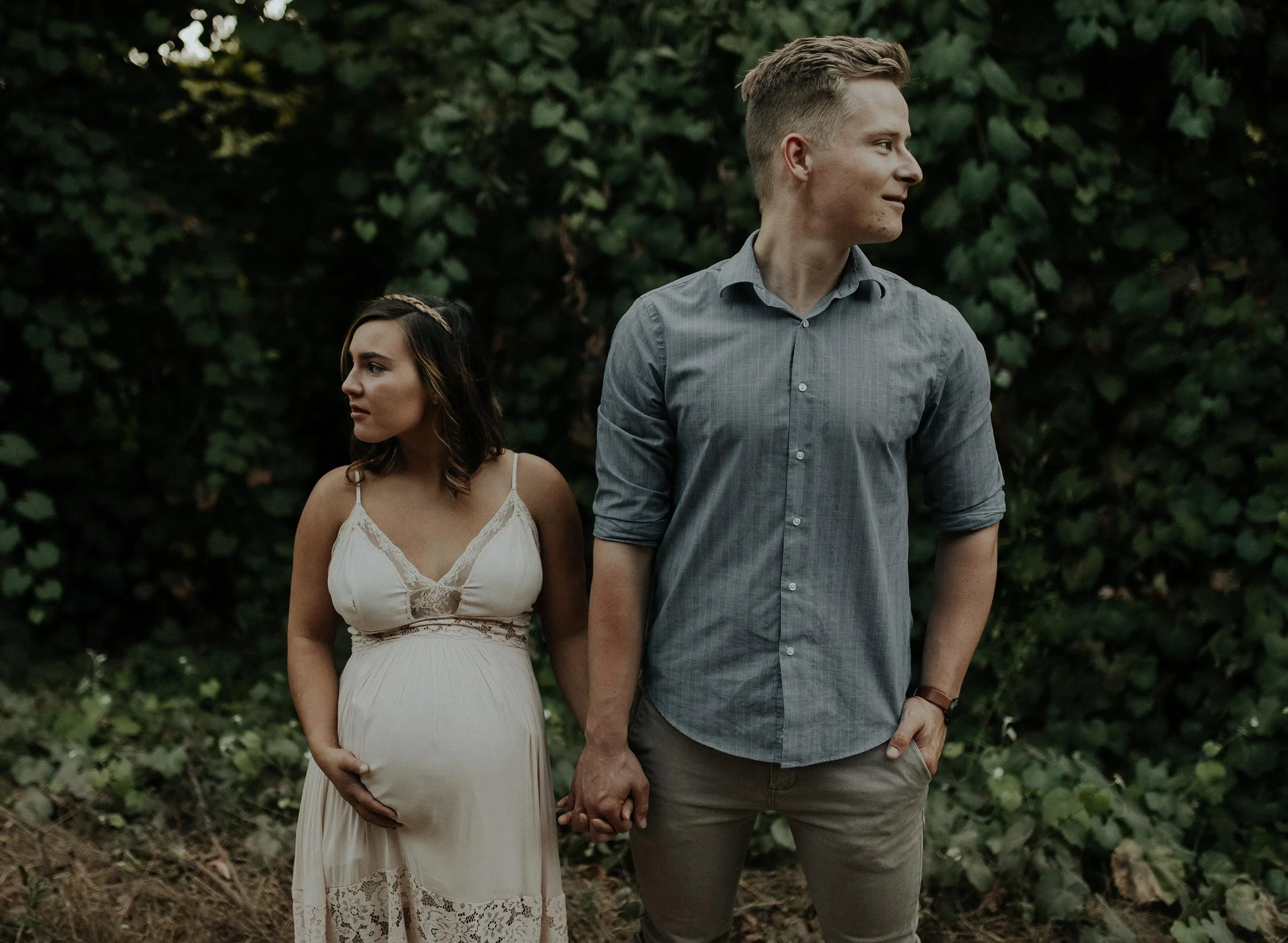 A pregnant woman wearing a white lace dress holds her belly while standing outside, holding hands with a man in a gray shirt and khaki pants, both standing against a background of green foliage.