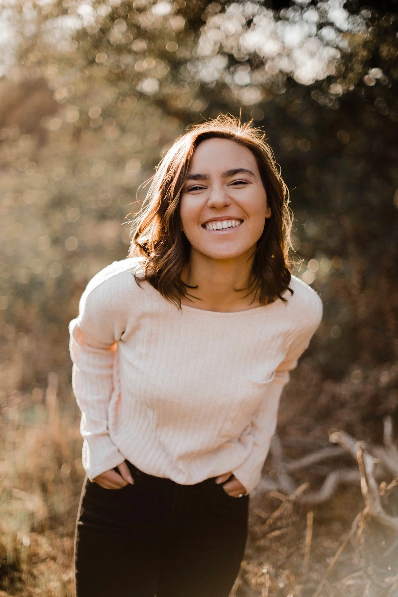 A woman with shoulder-length brown hair smiling, wearing a light-colored sweater and black pants, standing outdoors with blurred trees and branches in the background.