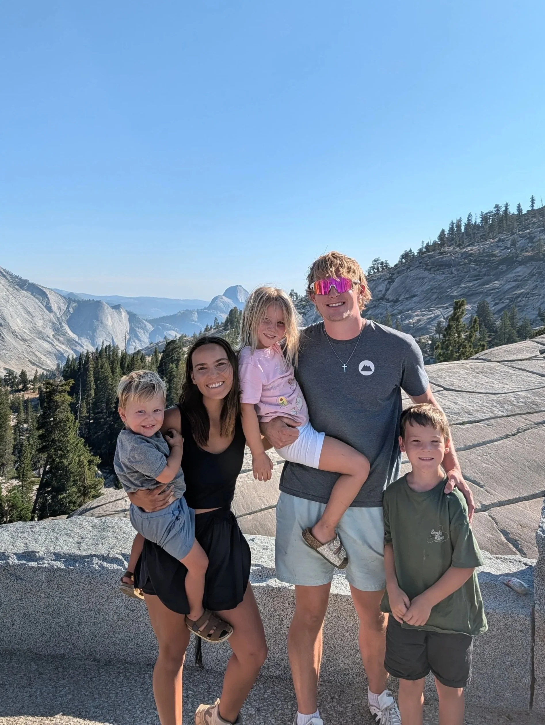 A group of five children and one adult posing together outdoors with a mountain landscape in the background. The sky is clear and blue.
