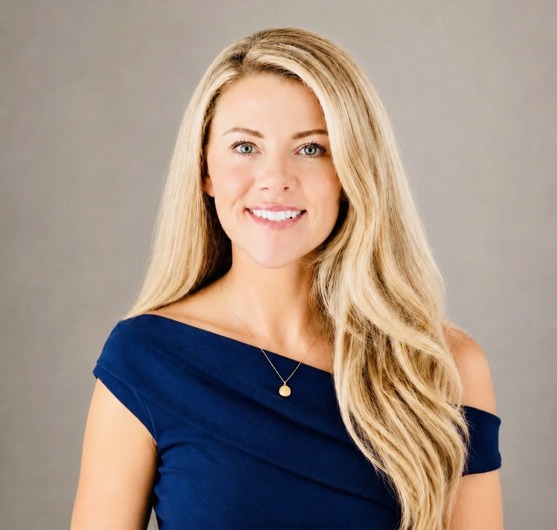 A woman with long blonde hair wearing a navy blue dress and a gold necklace, smiling against a plain gray background.