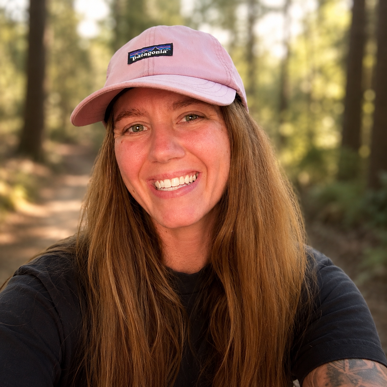 A smiling woman with long red hair wearing a pink Patagonia cap and black shirt, taking a selfie outdoors in a forested area with trees in the background.