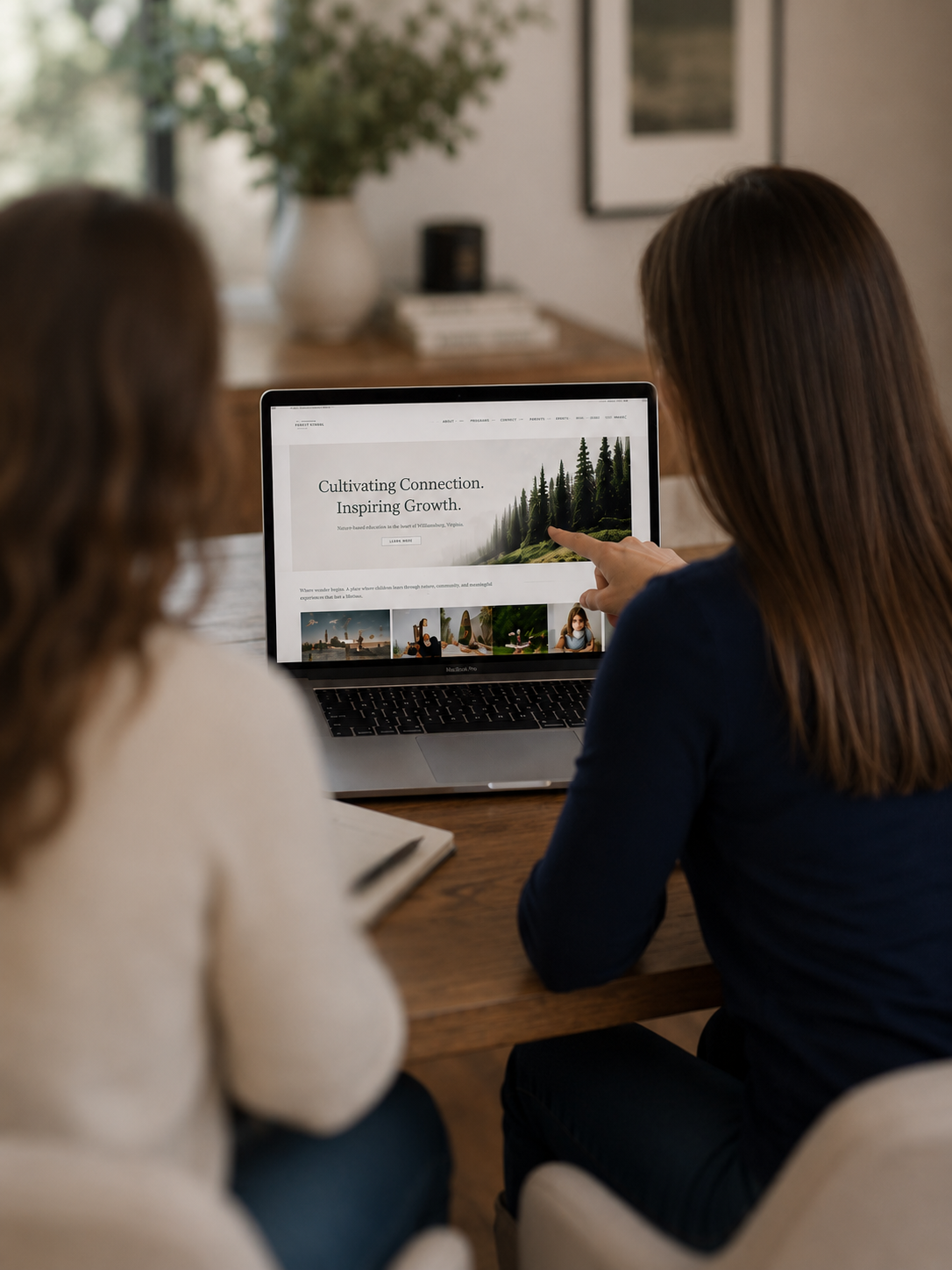 Two women sitting at a wooden table, one pointing at a website on a laptop, with a background of a blurred plant and framed pictures on the wall.